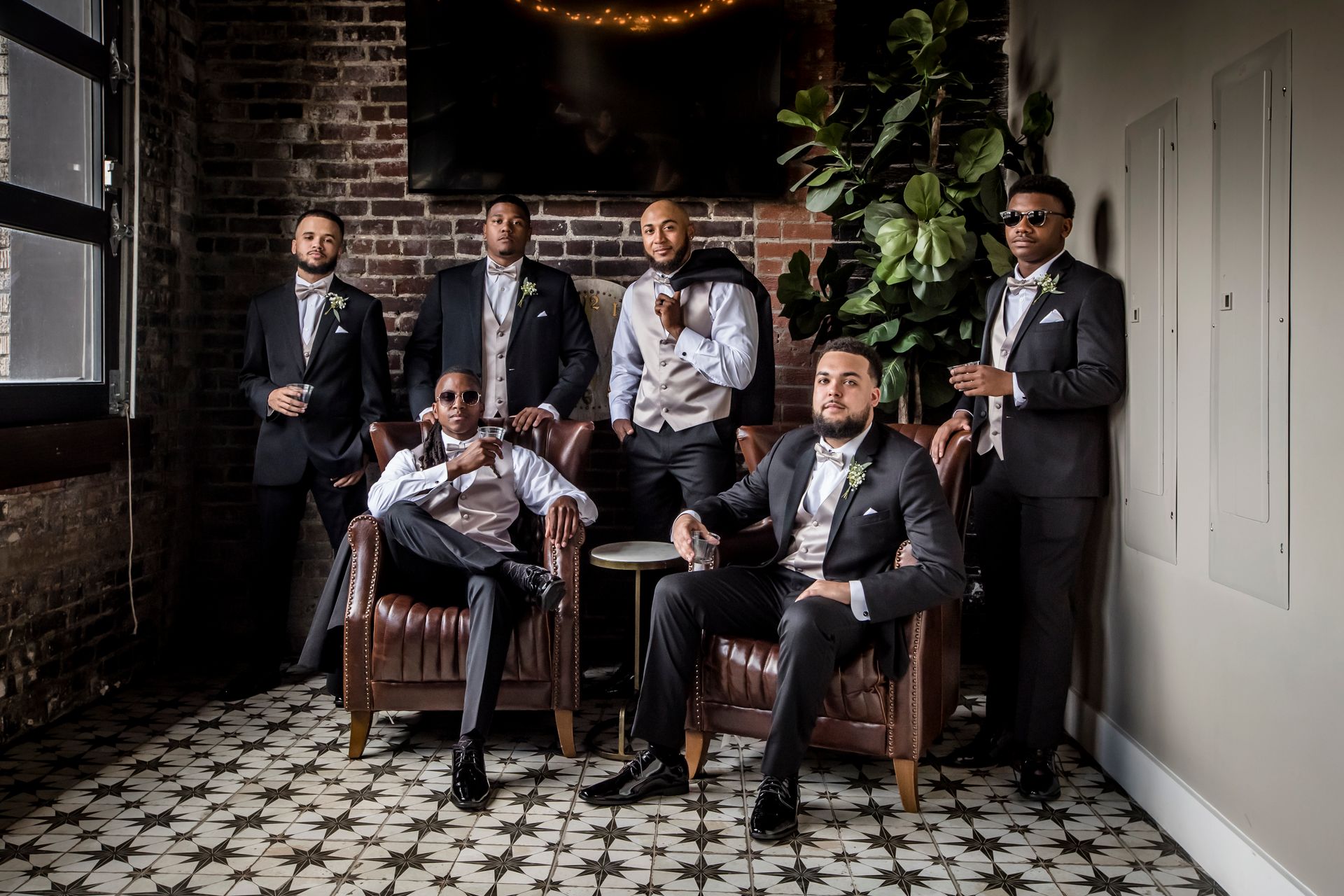 Five men in suits posing in a dimly lit, vintage-style room with patterned tile floor and plants