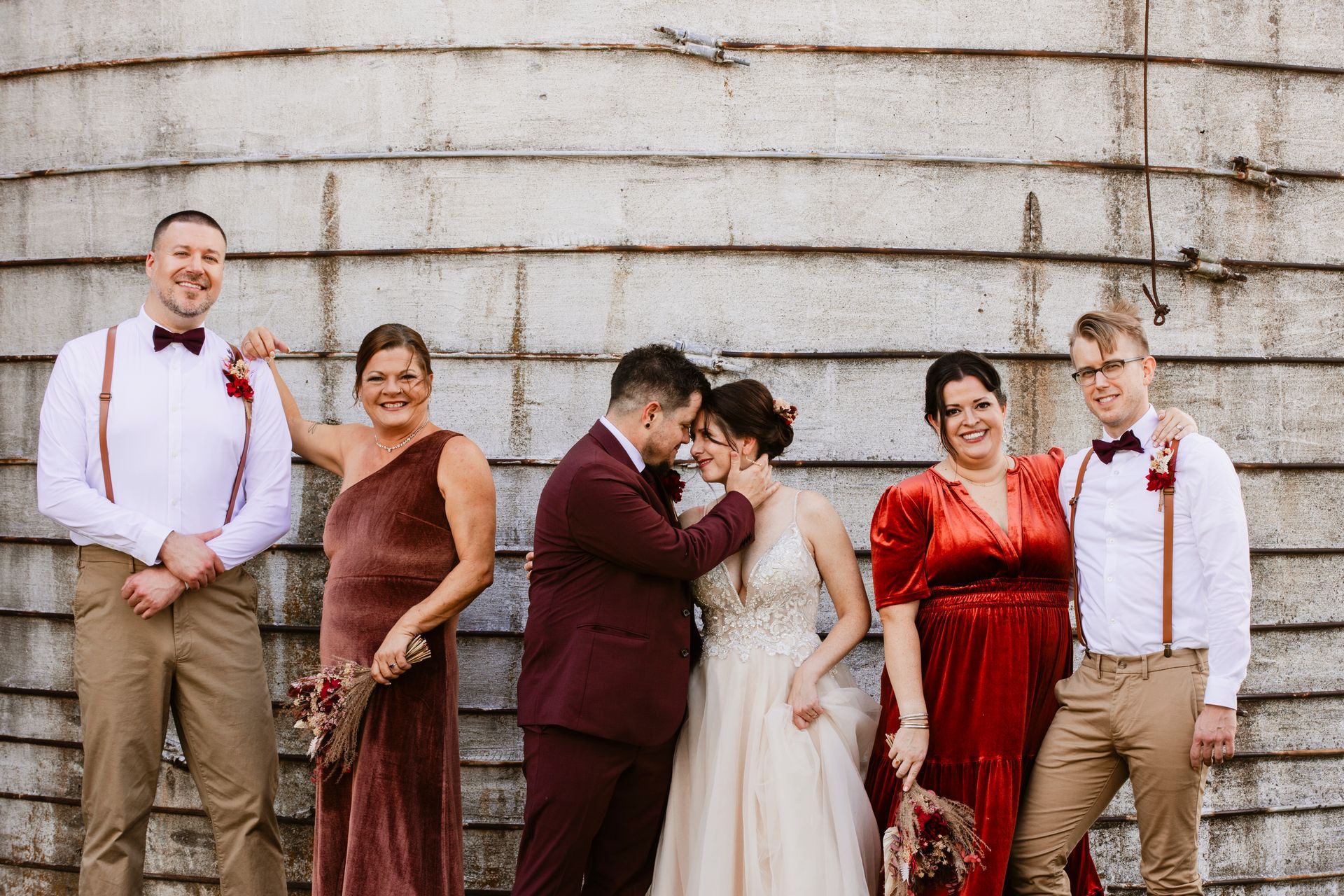 Wedding party posing in formal attire, smiling in front of a large stone wall.