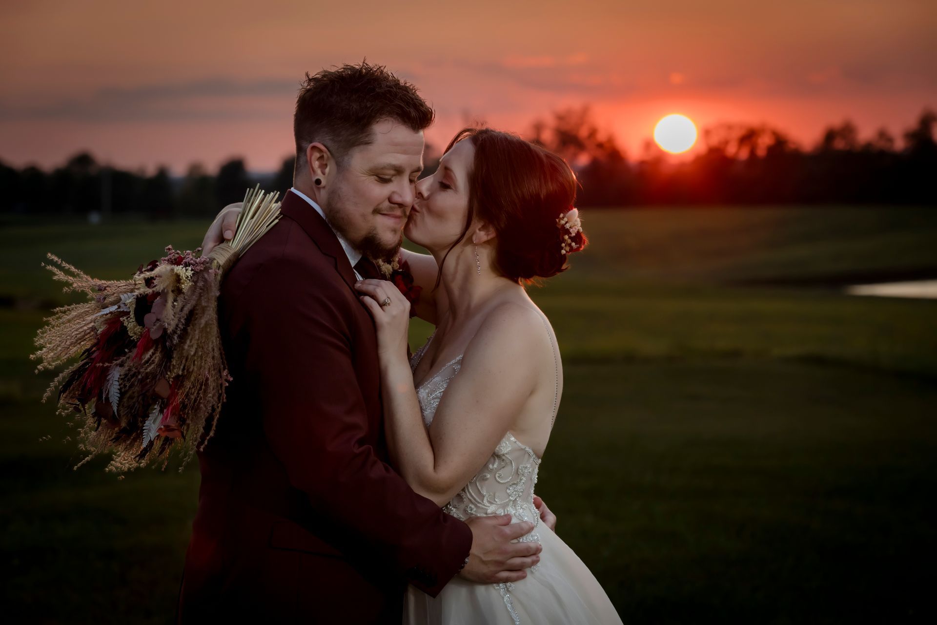 Couple embracing at sunset in a field, with warm orange sky and a bouquet of dried flowers