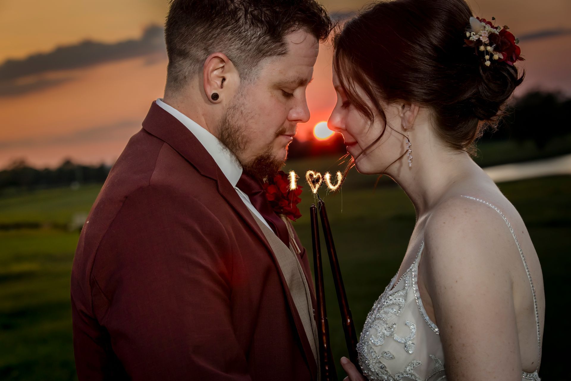 Couple in wedding attire touching foreheads at sunset in a field, with warm sunset light behind them