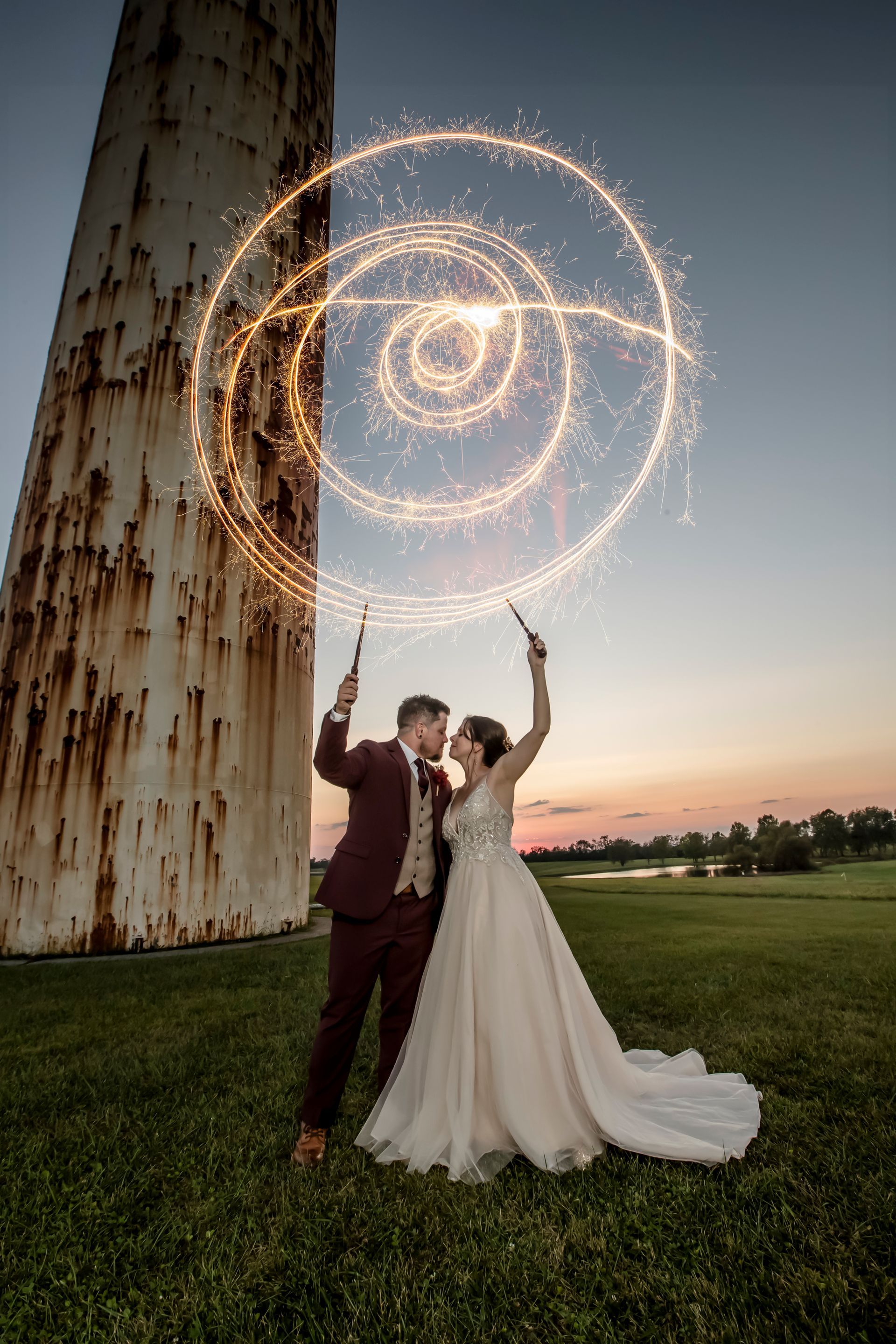 Bride and groom under a glowing spiral sparkler display on a lawn at sunset