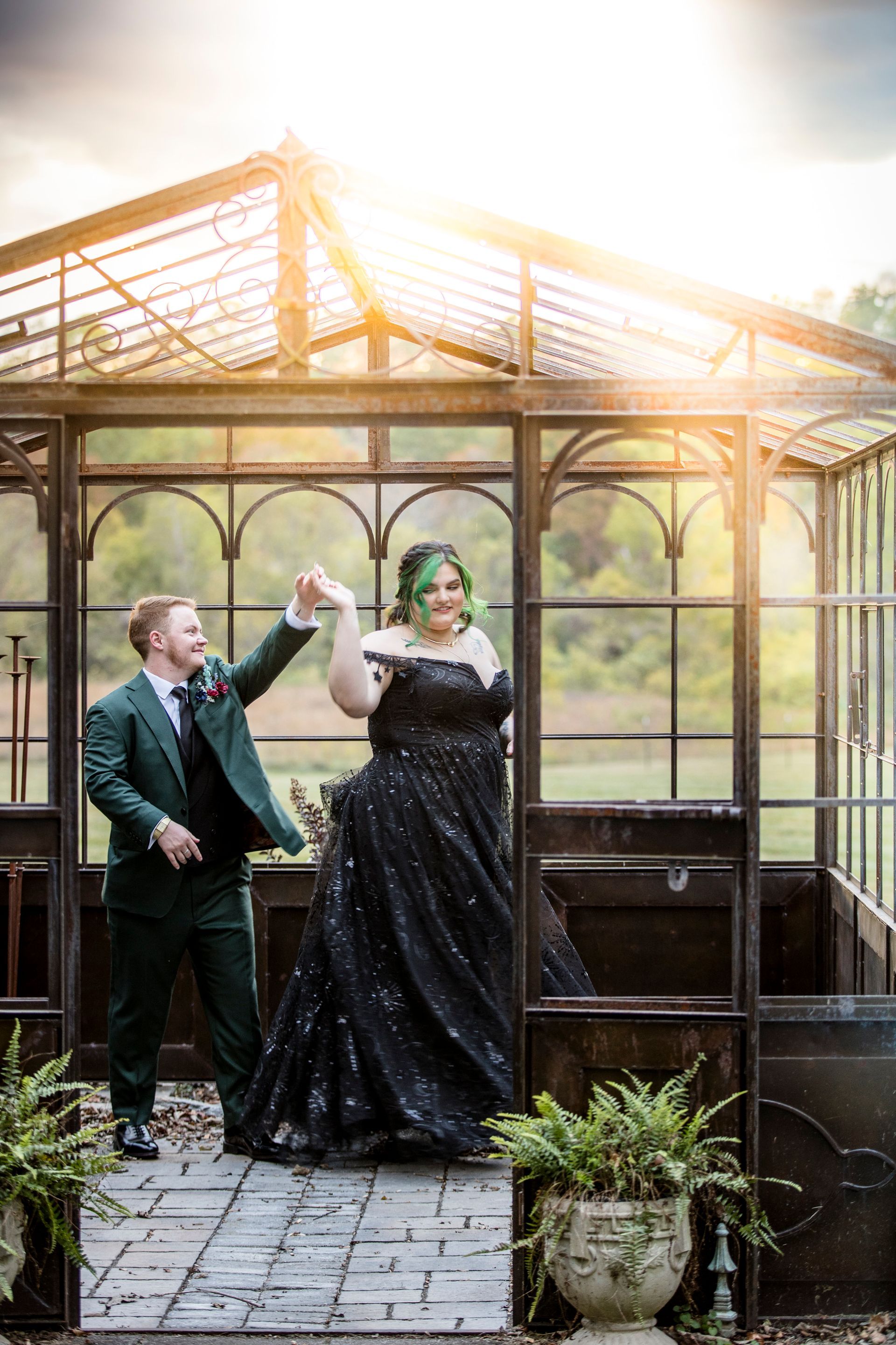 Couple in formal attire stand in a glass gazebo, raising hands beneath warm sunlight