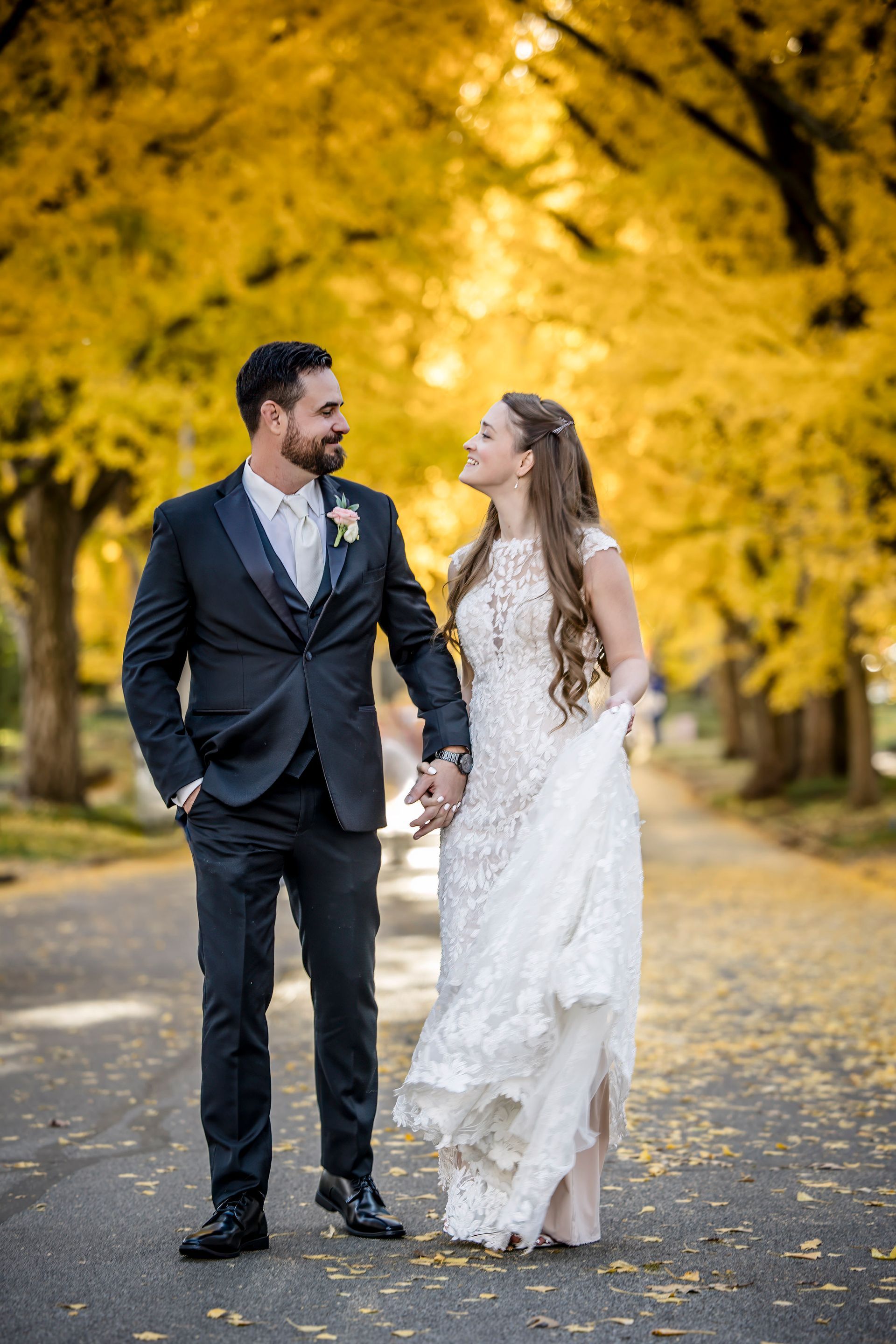 Couple in formal attire walking hand in hand on a tree-lined path with golden autumn leaves.