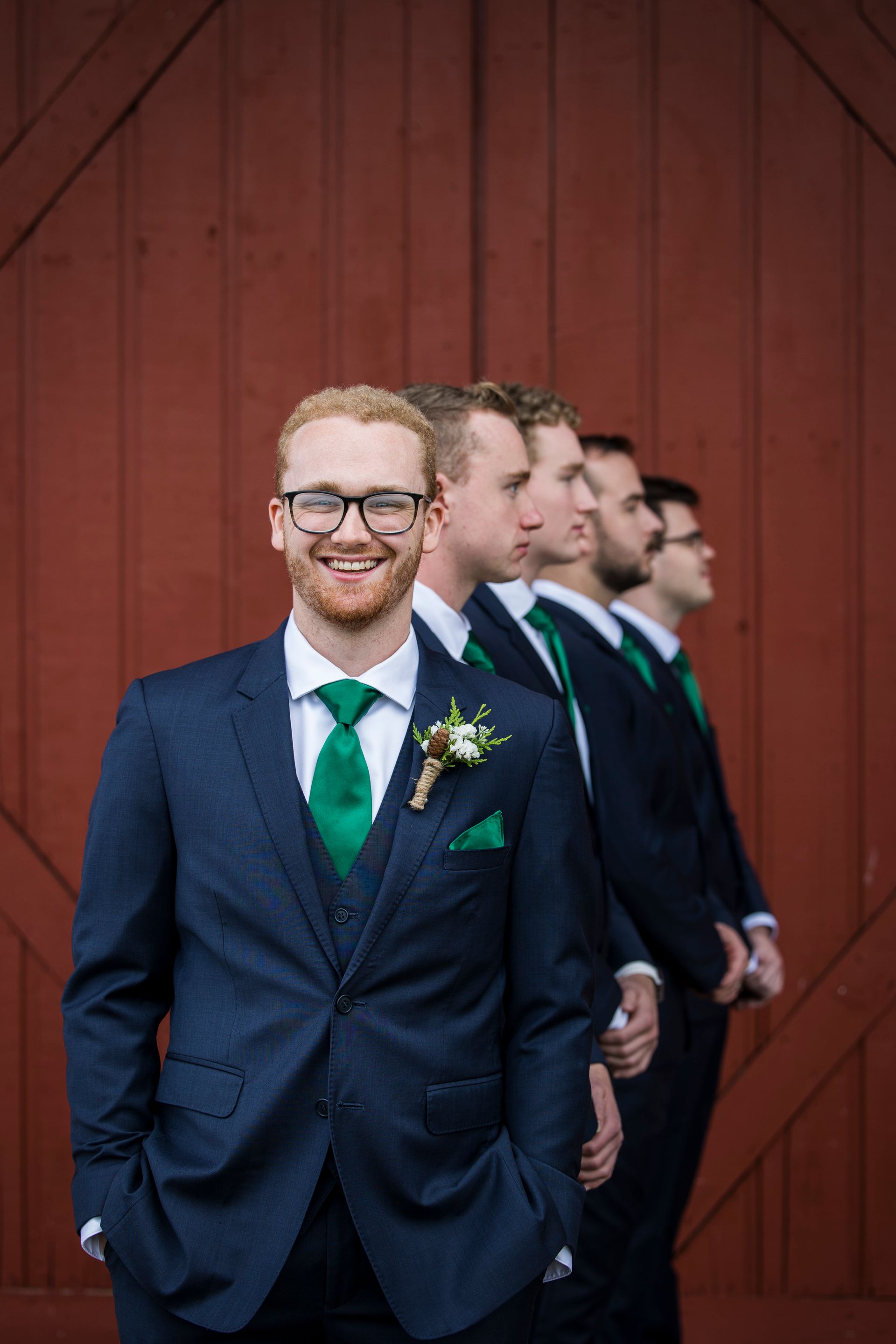 Four men in navy suits and green ties standing in a line in front of a red wooden wall.