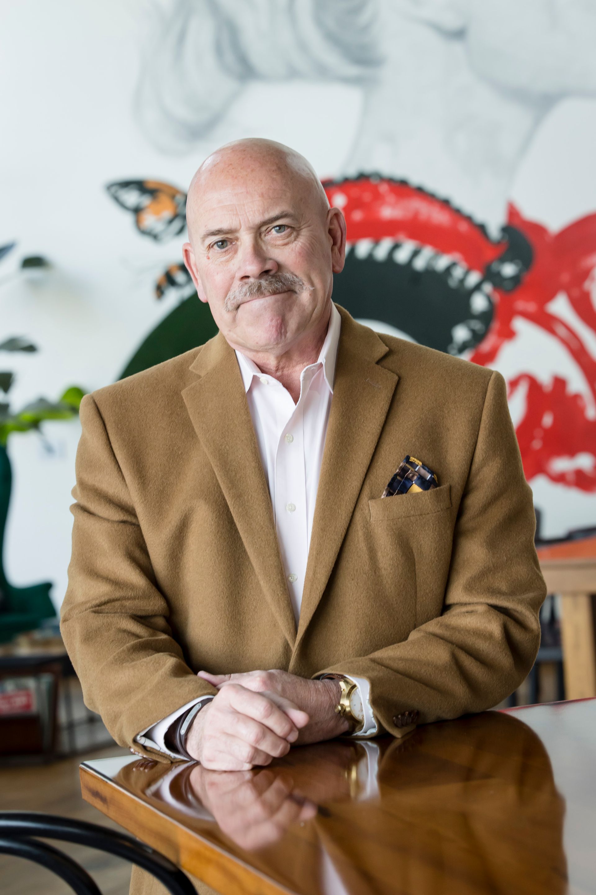Man in a tan blazer stands by a polished table in front of a colorful butterfly mural.