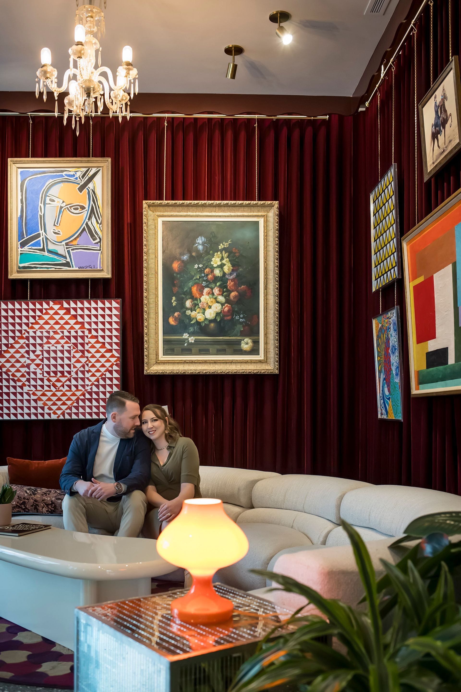 Couple seated on a sofa in a richly decorated lounge with framed art, red curtains, and warm lamps.