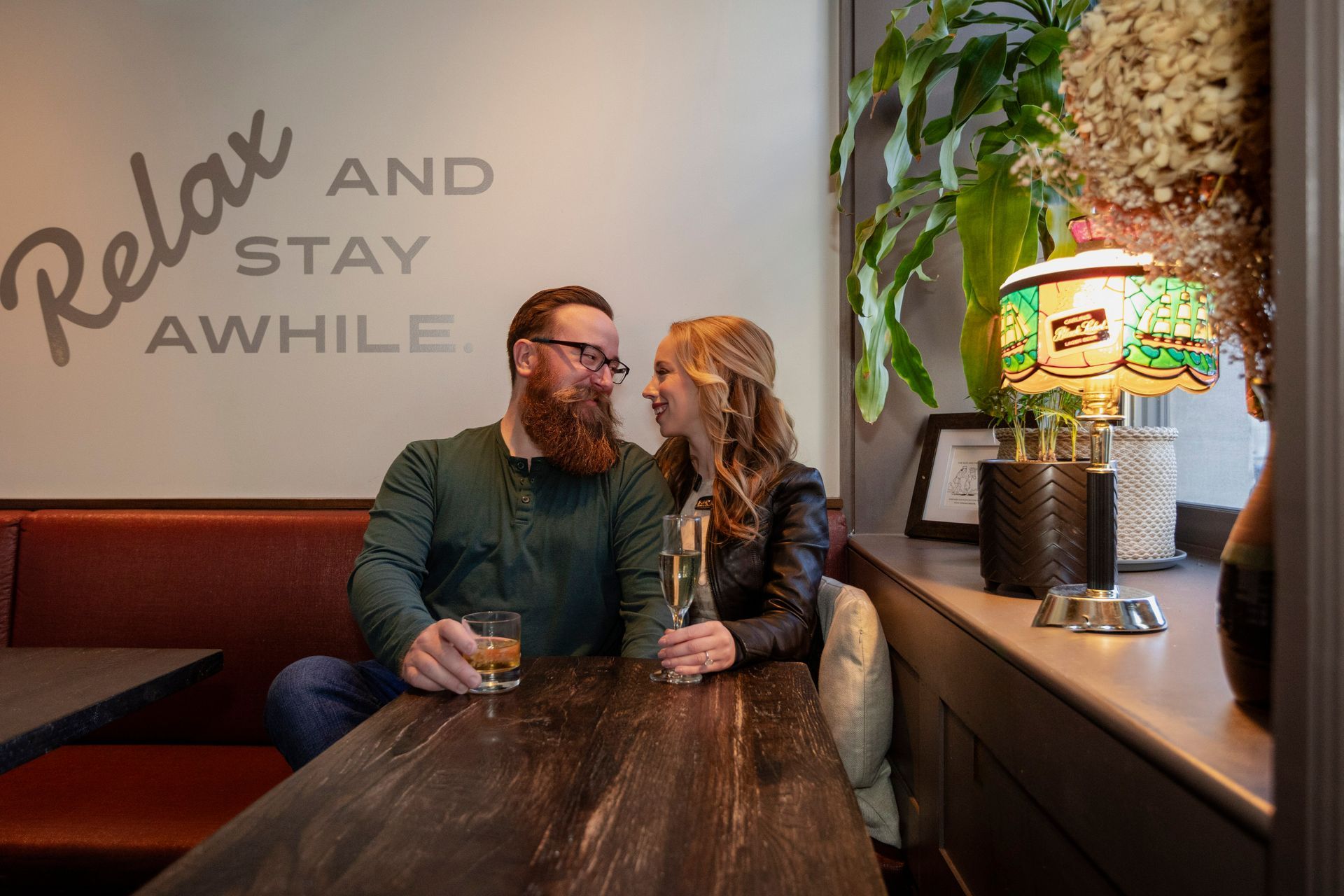Two people seated at a cozy bar booth, smiling at each other beside a colorful lamp and wall sign.