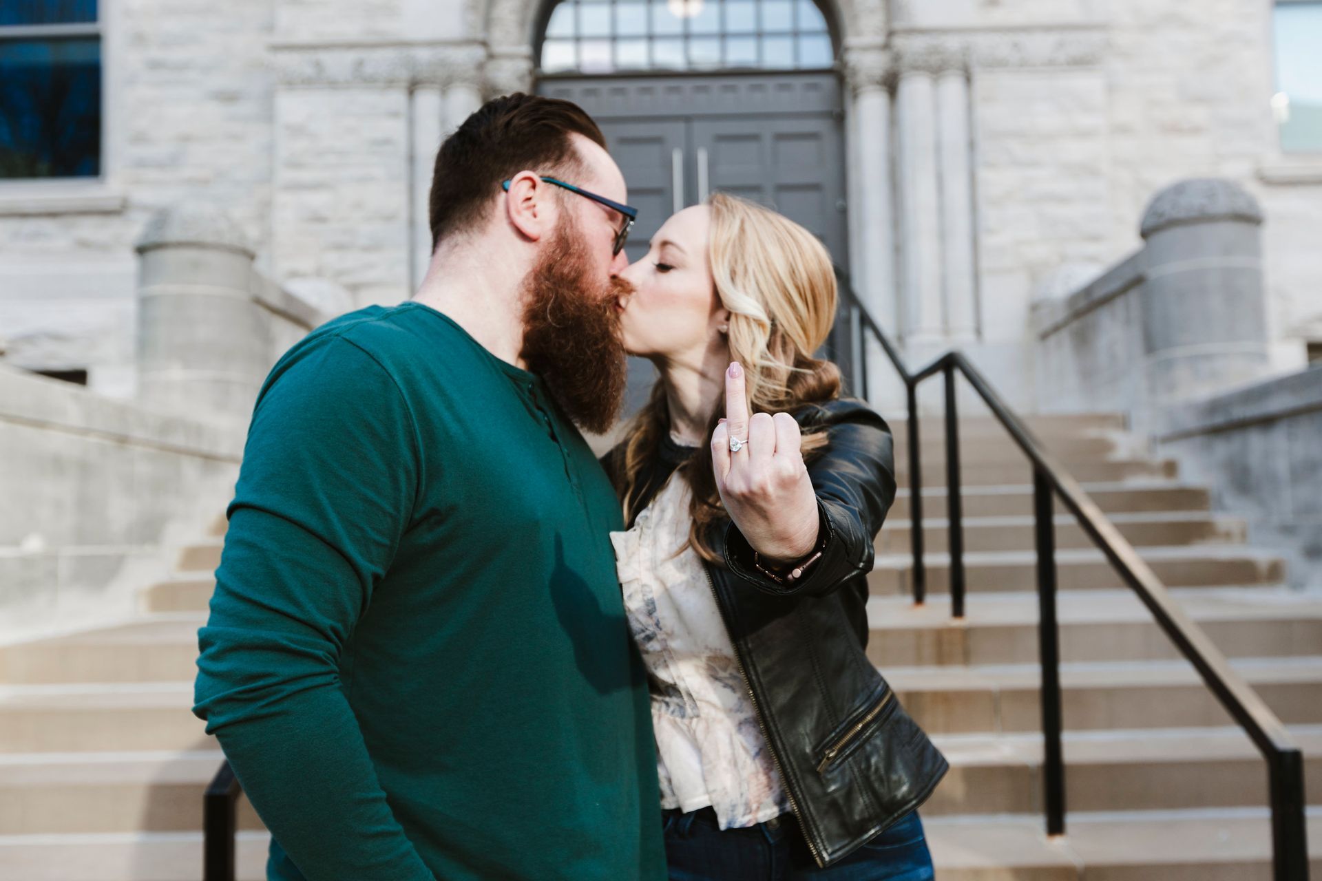 Couple kissing on stone steps outside a building, the man in a teal shirt and the woman in a black jacket.