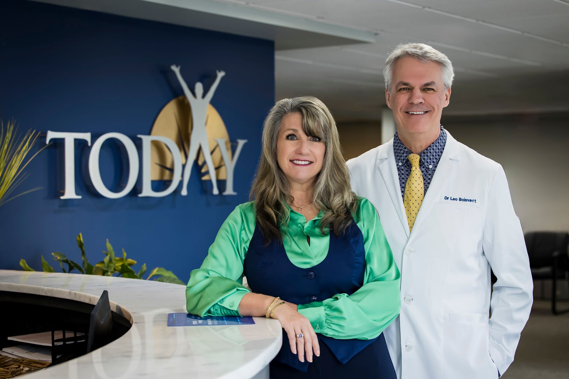Two smiling staff members stand in a clinic lobby beside a “TODAY” sign.