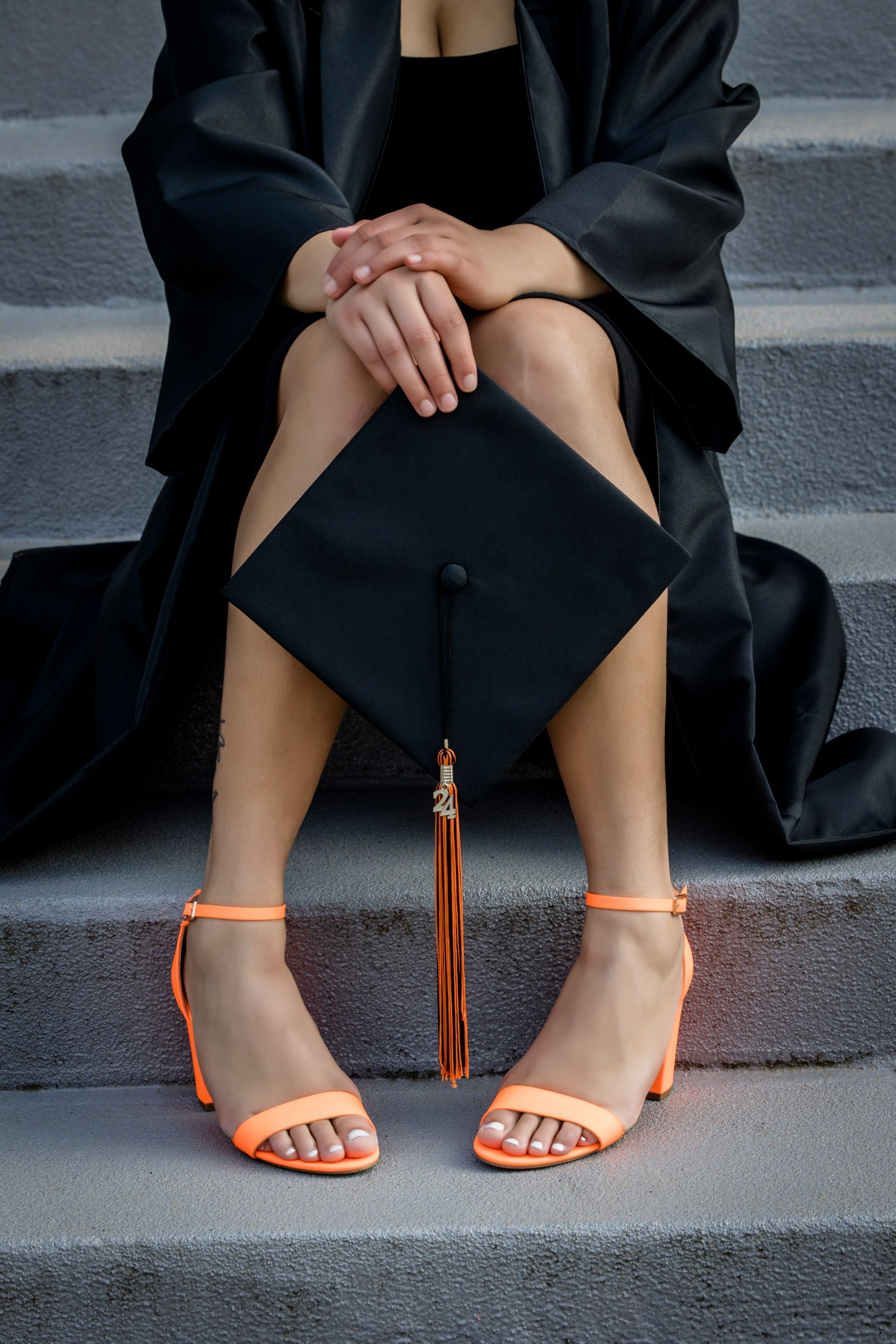 Person sitting on steps with black graduation cap on lap, wearing orange-strapped heels.