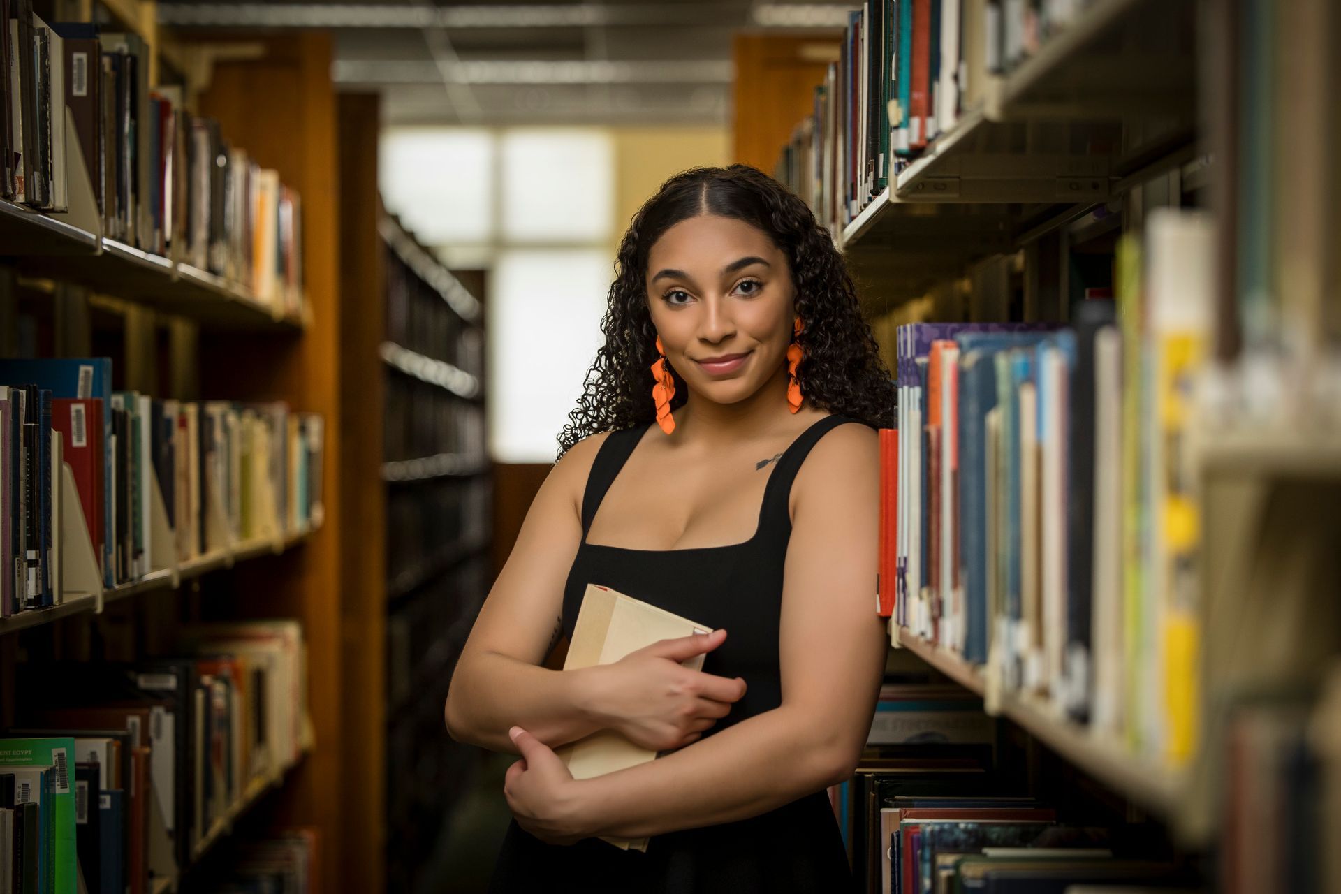 Woman holding a notebook in a library aisle, standing between bookshelves and facing the camera.