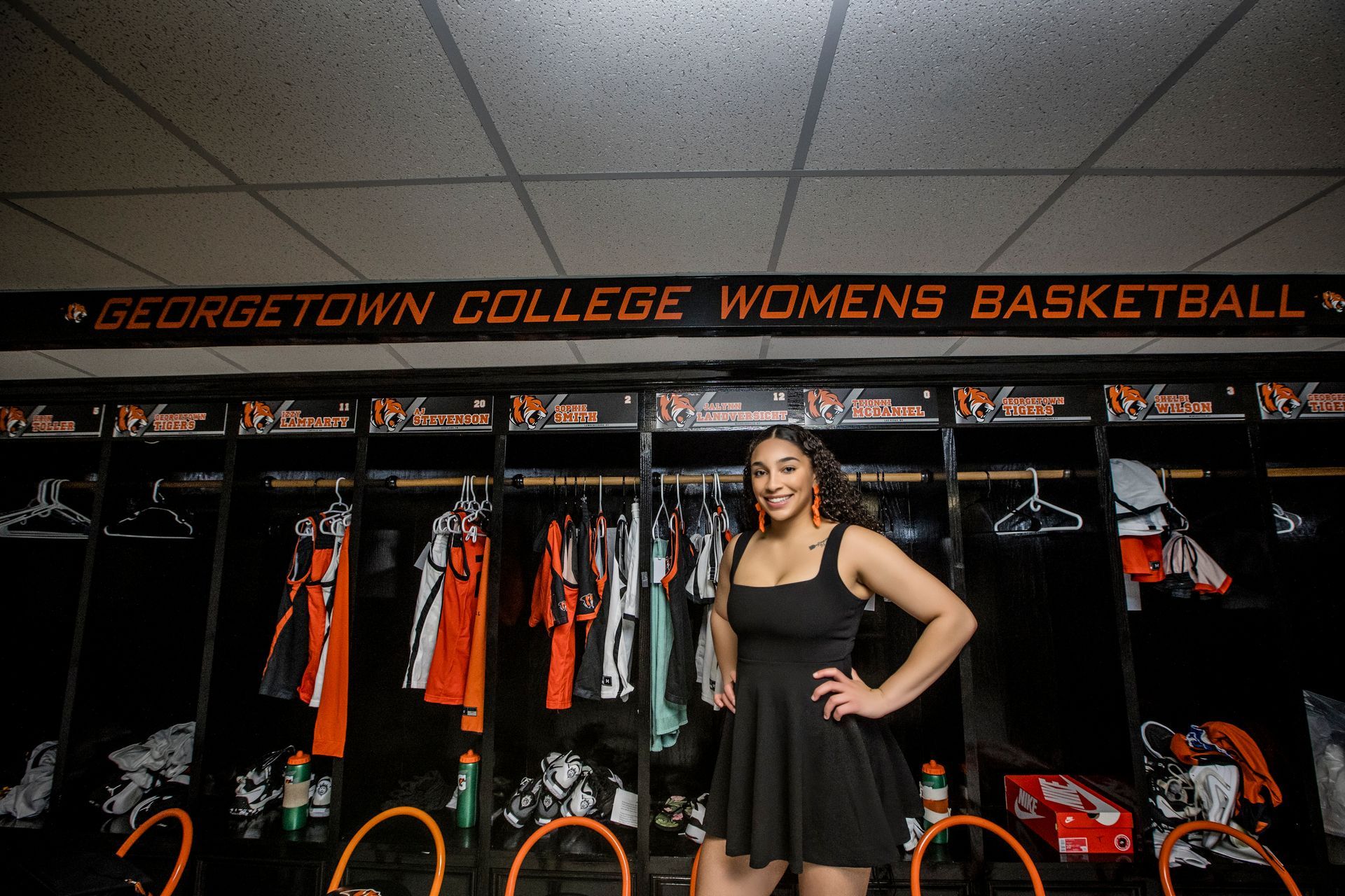 Georgetown College women’s basketball locker room with a woman in a black dress posing in front of orange-and-black lockers