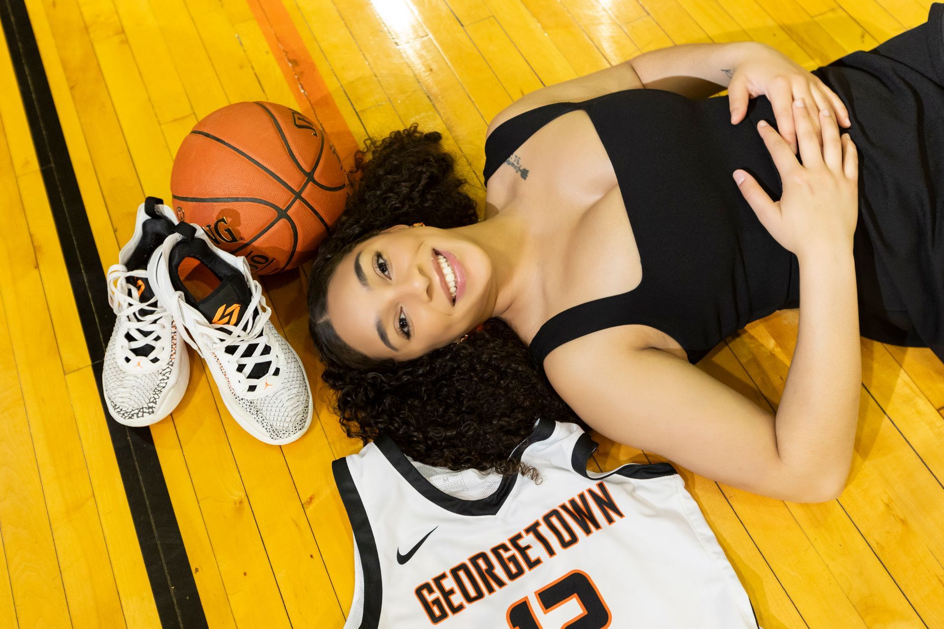 Smiling basketball player lying on gym floor beside shoes, ball, and Georgetown jersey