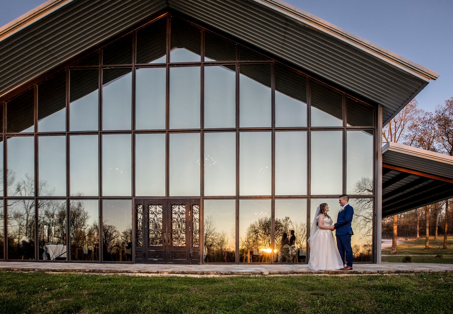 Two people in formal attire stand outside a glass-front building at sunset.