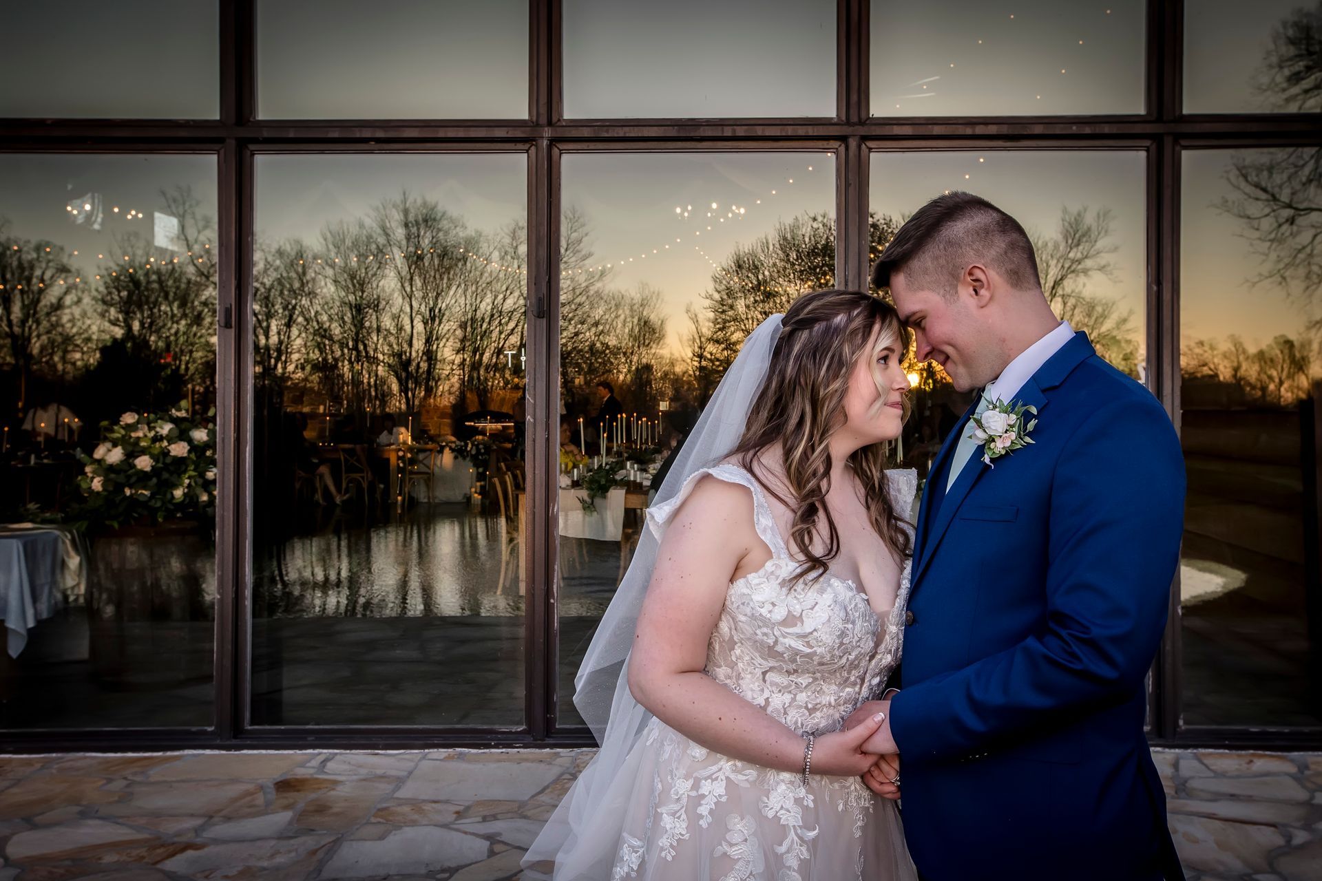 Bride and groom embracing in front of large windows at sunset, with reflections and string lights outside.