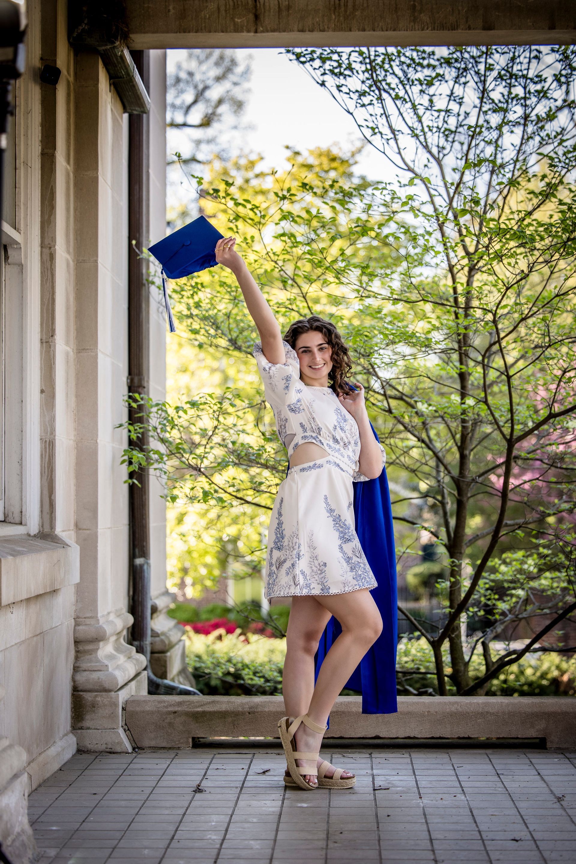 Woman in a white dress posing under a porch, holding a blue book overhead, with trees and flowers behind.