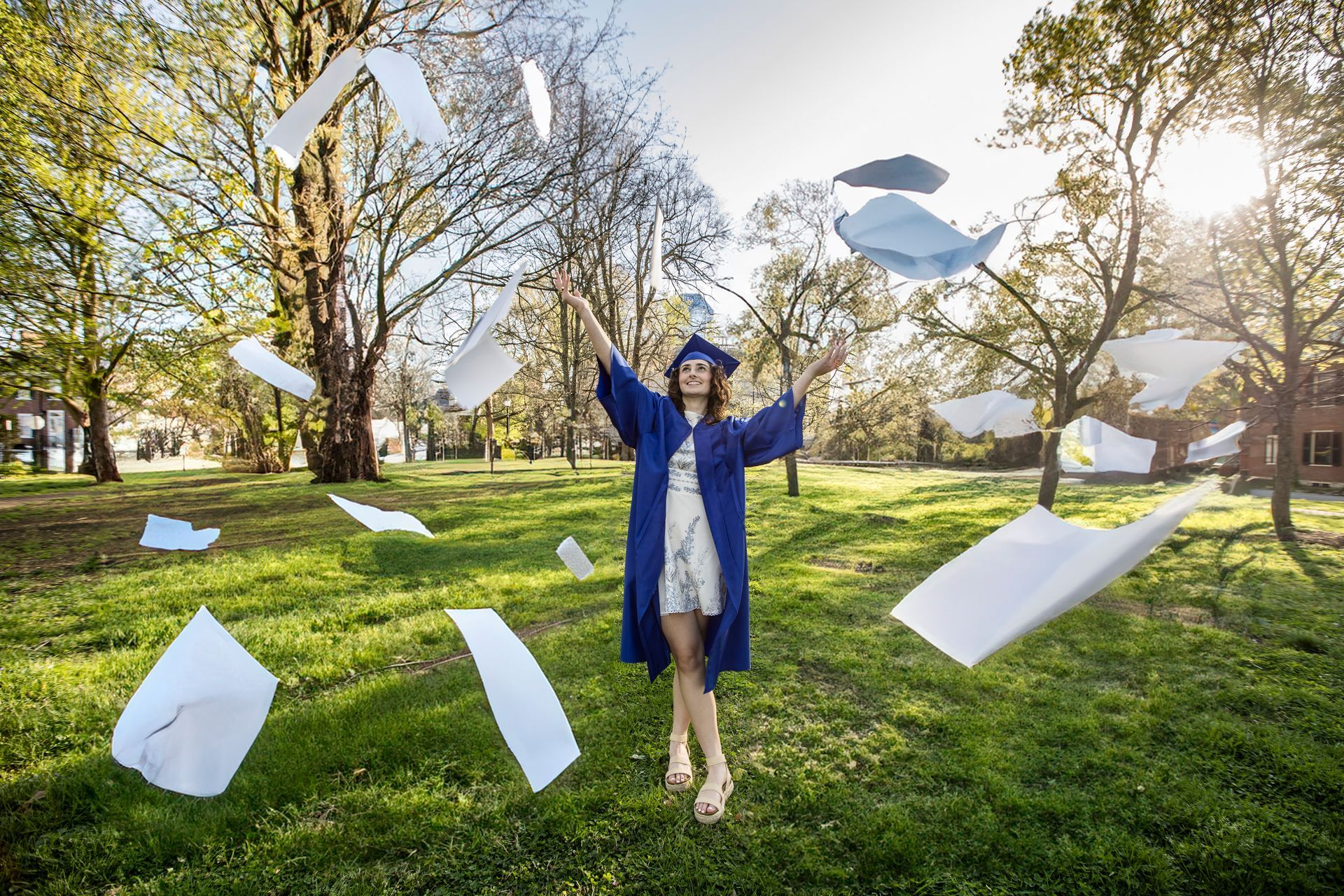 Graduate in blue gown tossing papers in a sunny park