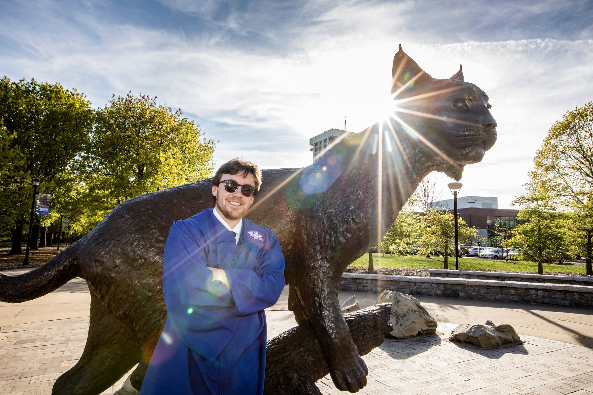 Person in blue jacket posing beside a large animal statue outdoors, with sun flare behind them.