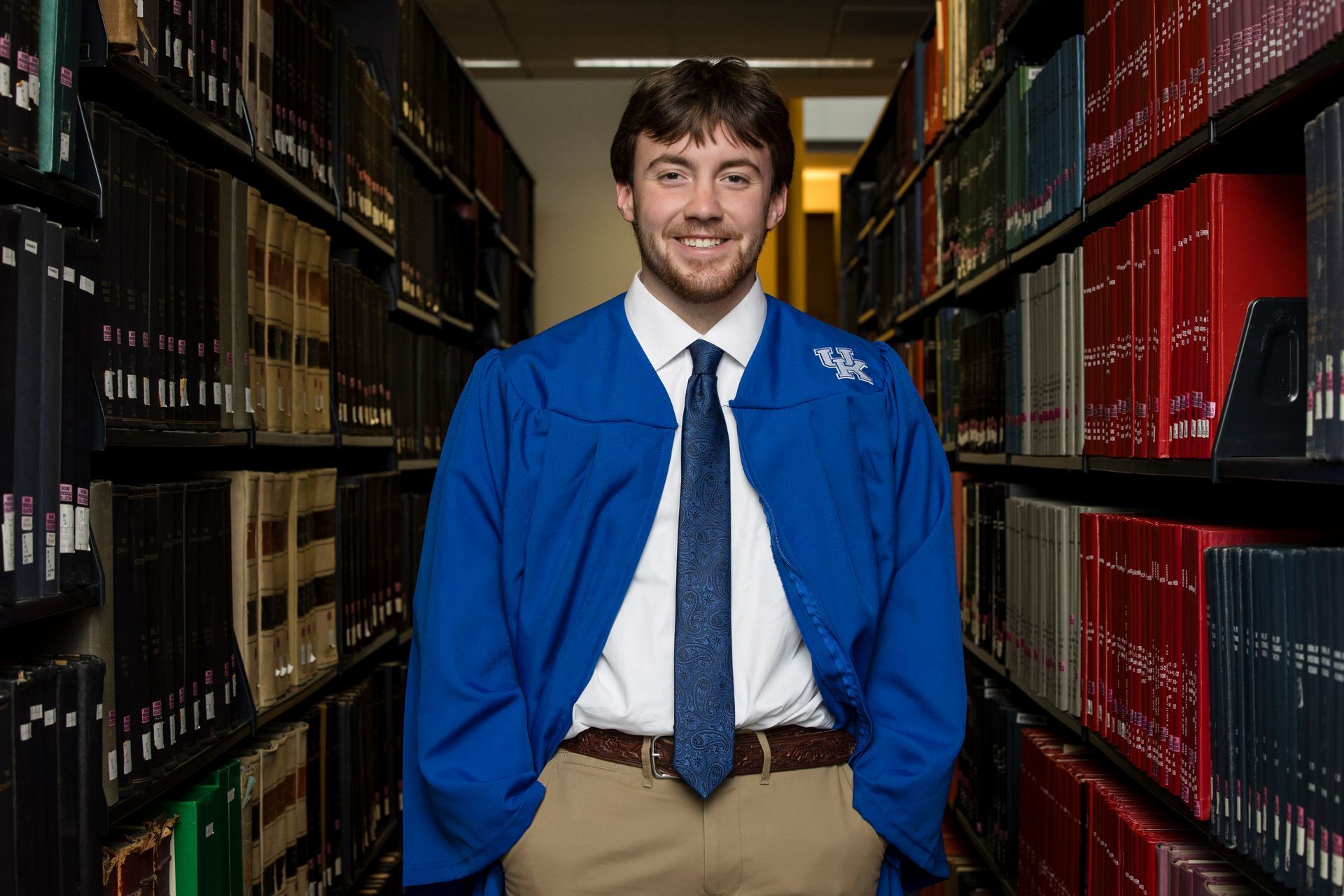 Young man in blue coat and tie standing between library shelves and smiling