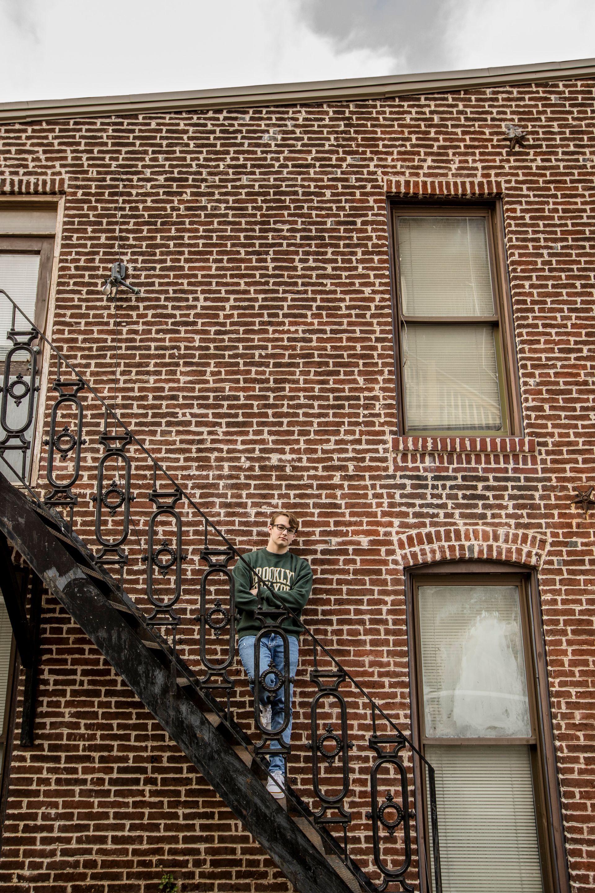 Brick building facade with metal fire escape and two windows, one with curtains, under an overcast sky