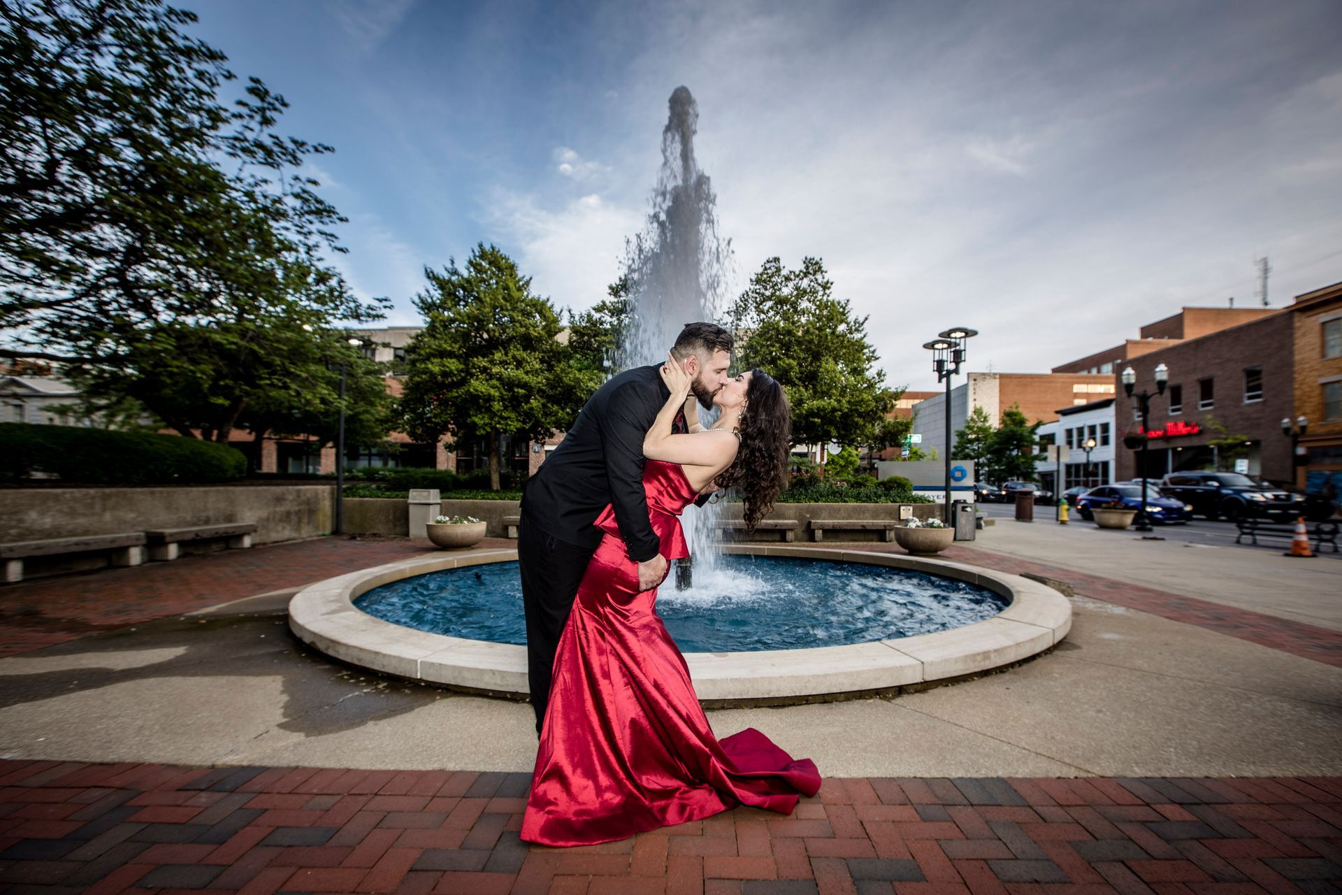 Couple in formal attire kissing by a fountain in a town square