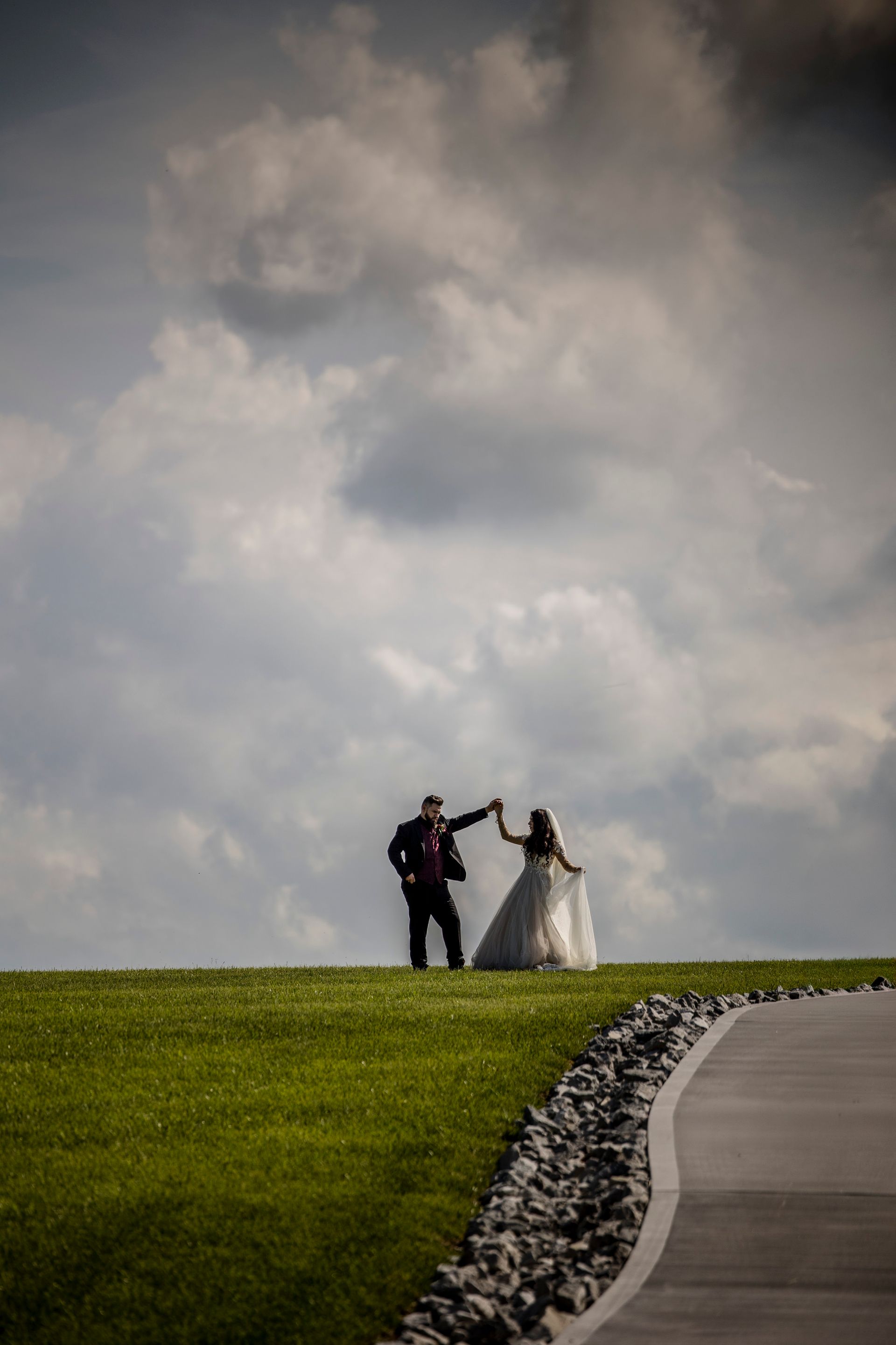 Wedding couple holding hands on a grassy hill under cloudy sky, beside a curving path