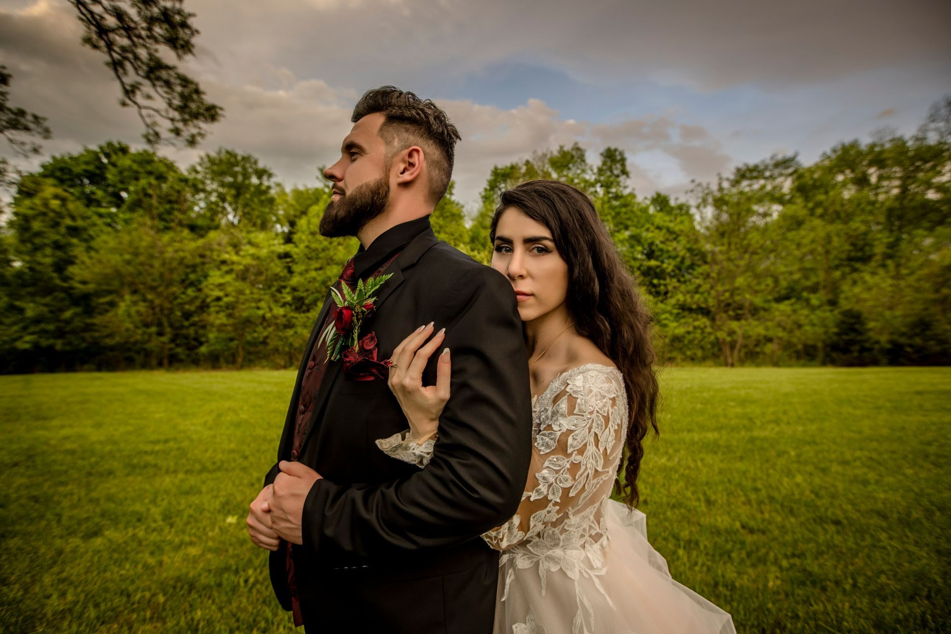 Couple in formal attire standing in a green field, with the woman leaning on the man under a cloudy sky.