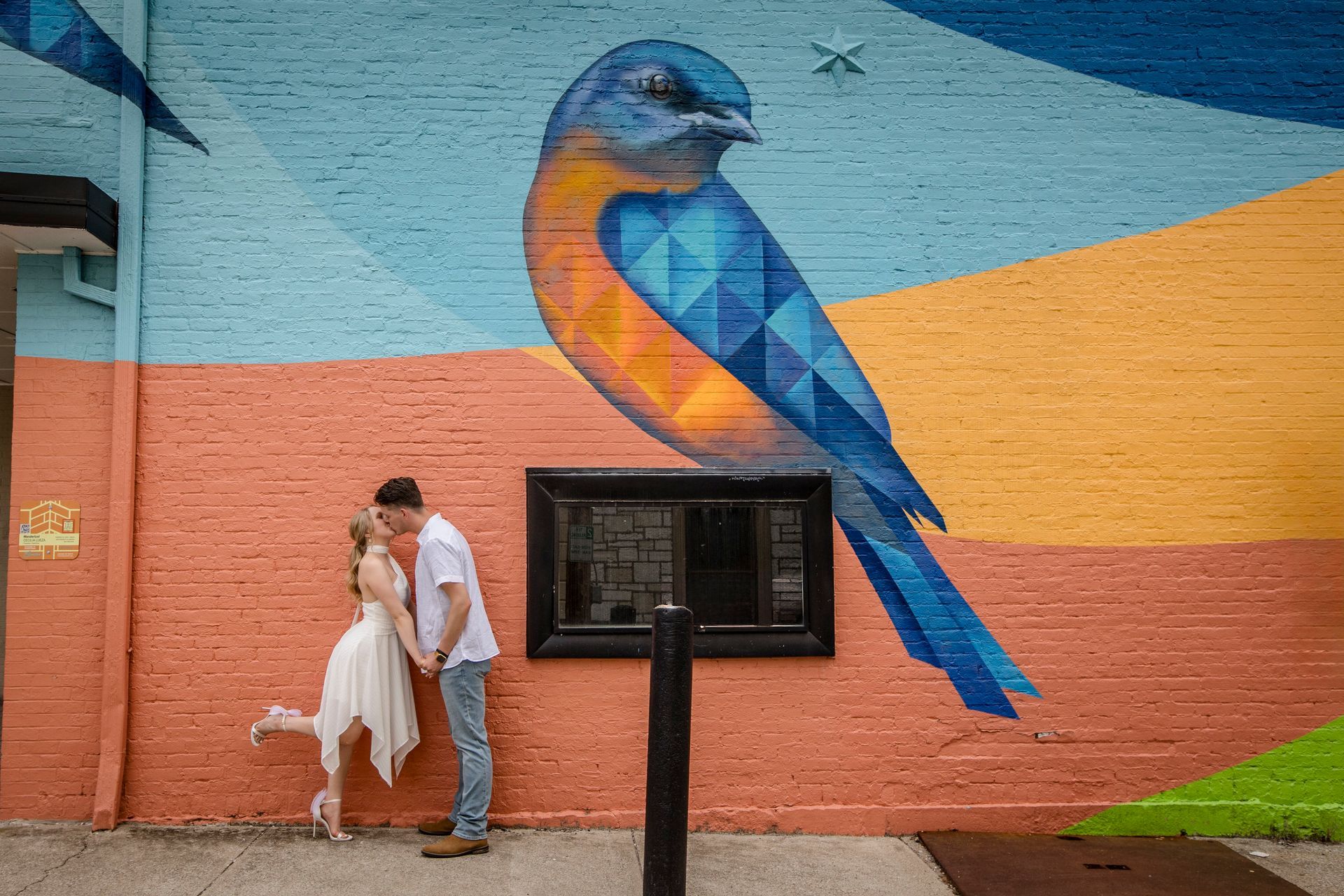 Couple embracing in front of colorful bird mural on a brick wall