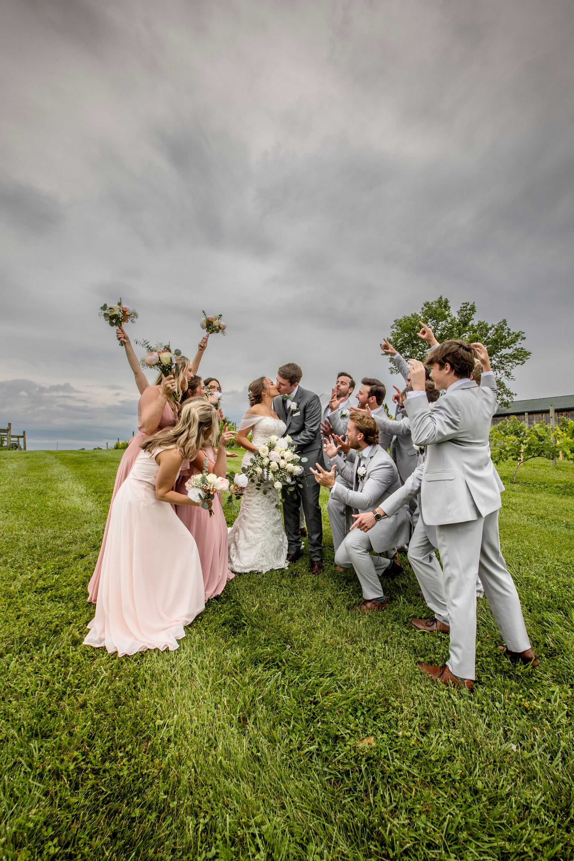 Wedding party posing on a grassy hill under a cloudy sky, with bouquets and light gray suits