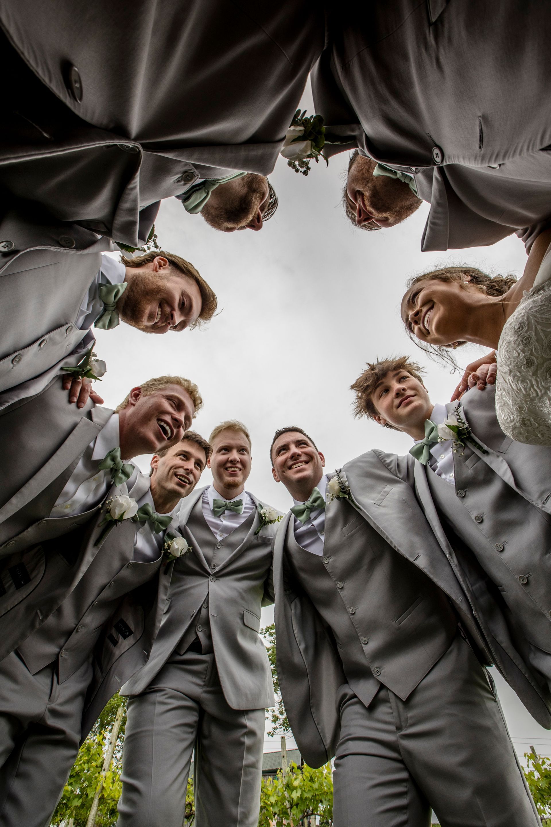 Groomsmen in gray suits lean in a circle, smiling down at the camera outdoors.