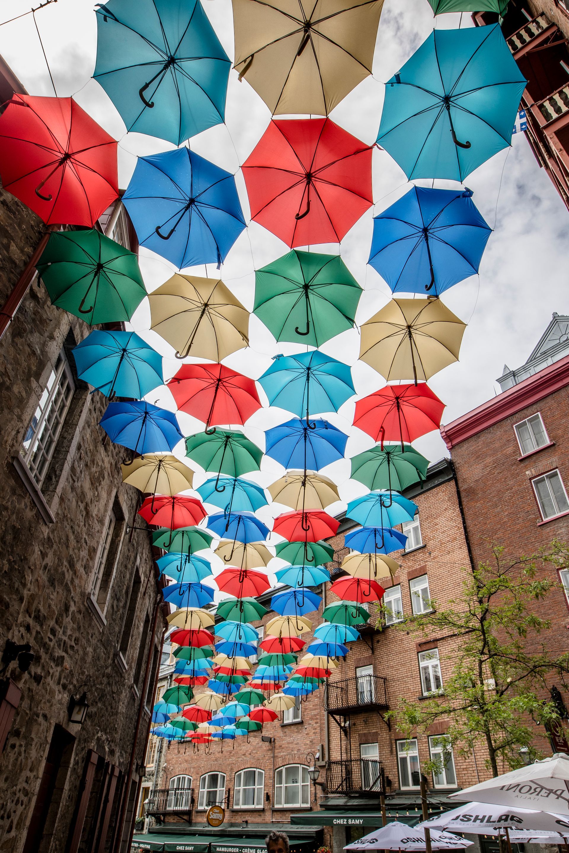 Colorful umbrellas hanging overhead in a narrow brick alley with buildings lining both sides.