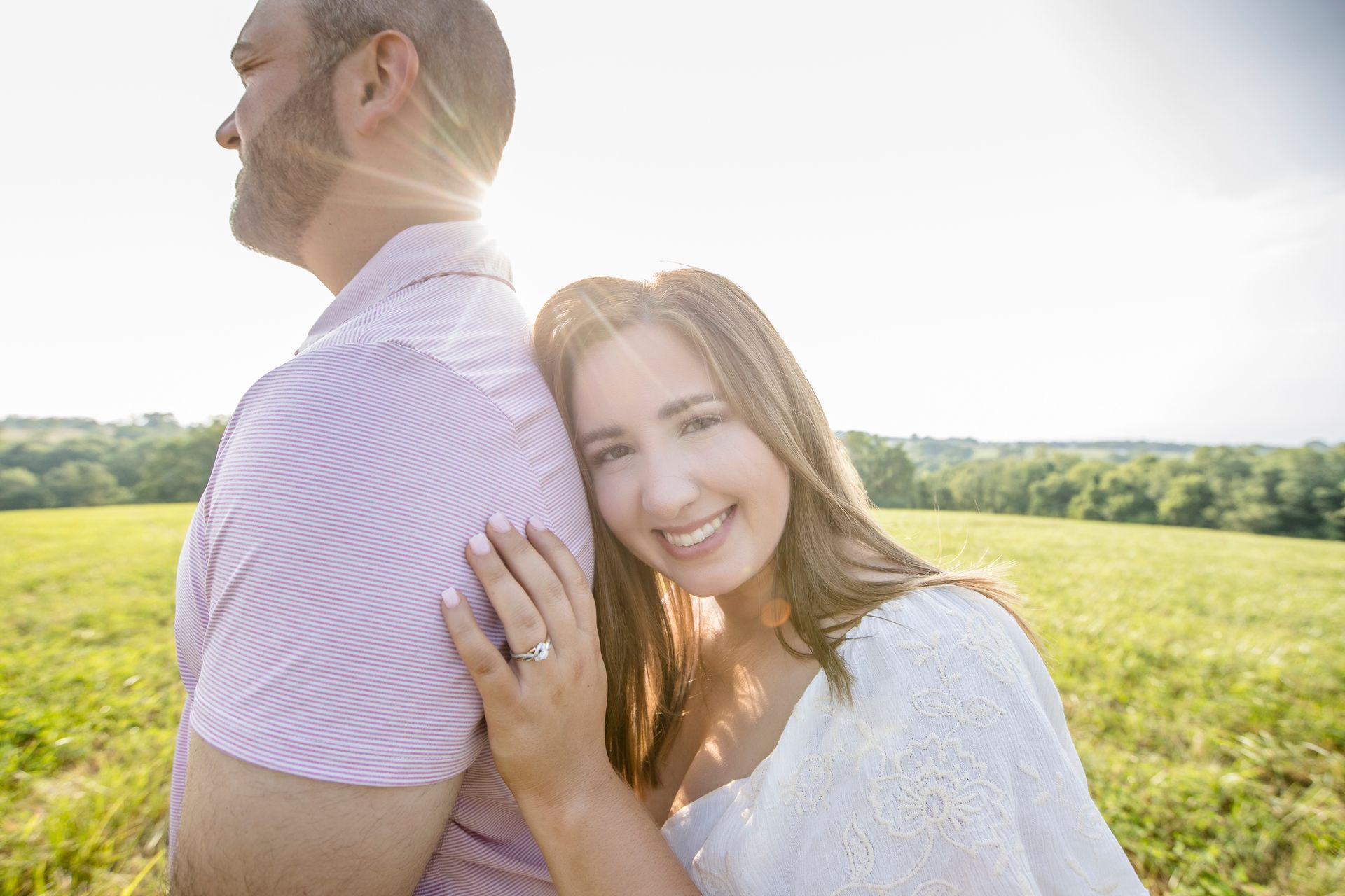 Couple embracing in a sunlit field, smiling with backlight and green hills behind them