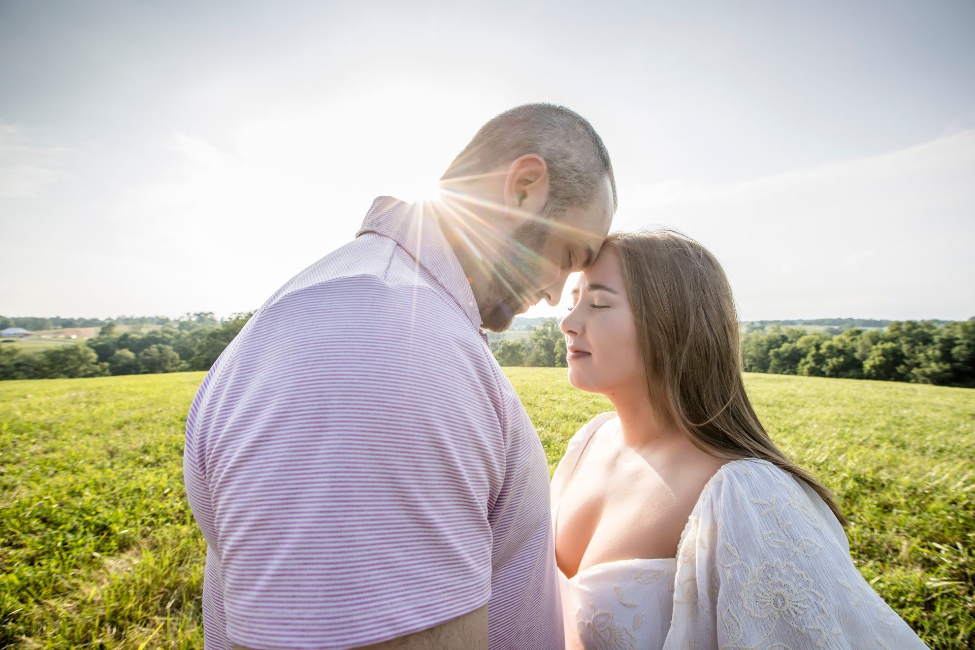 Couple standing close in a sunlit green field, facing each other with a bright sky behind them