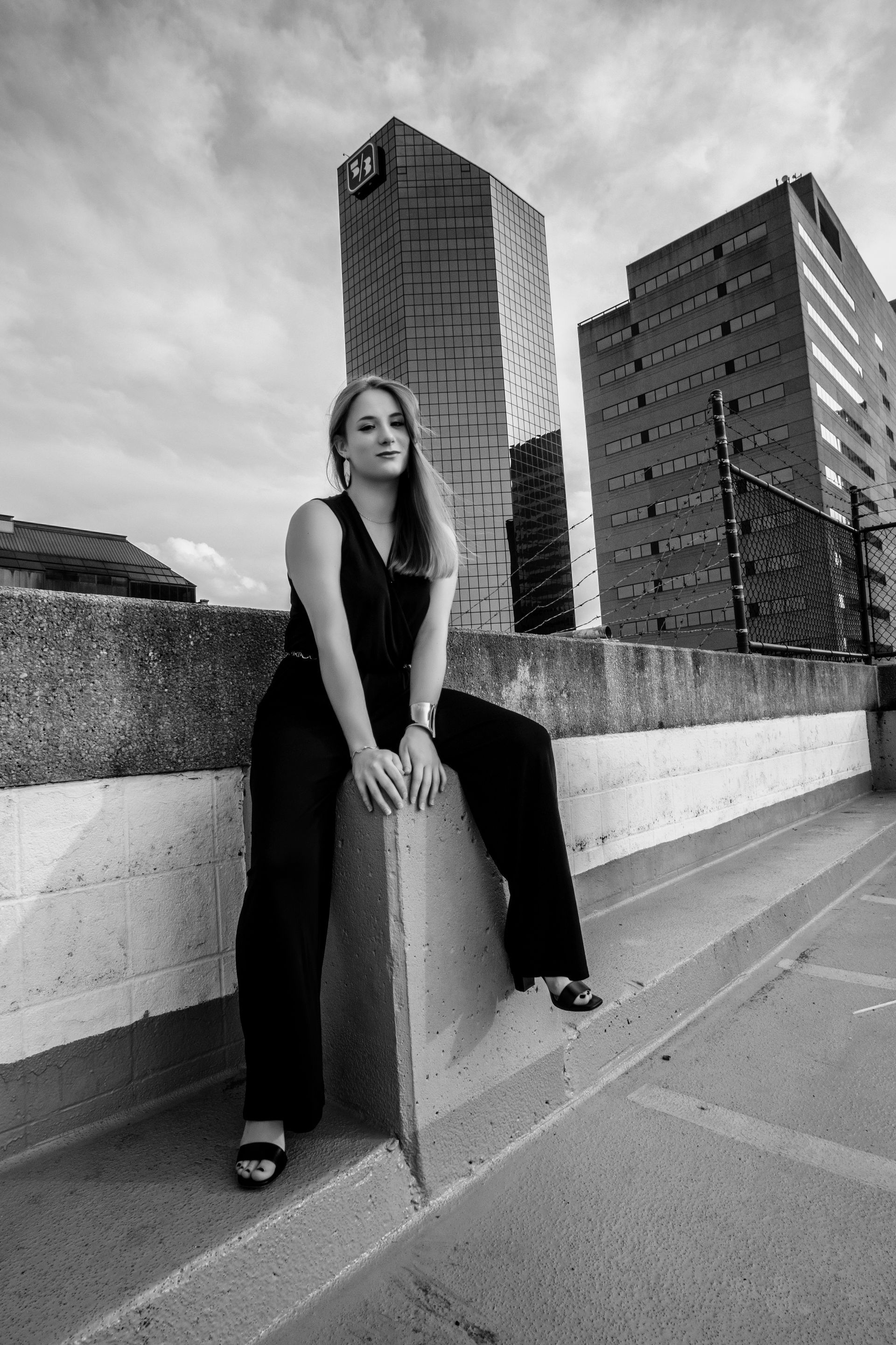 Person sitting on a concrete ledge in front of tall buildings, wearing dark clothing, black-and-white photo