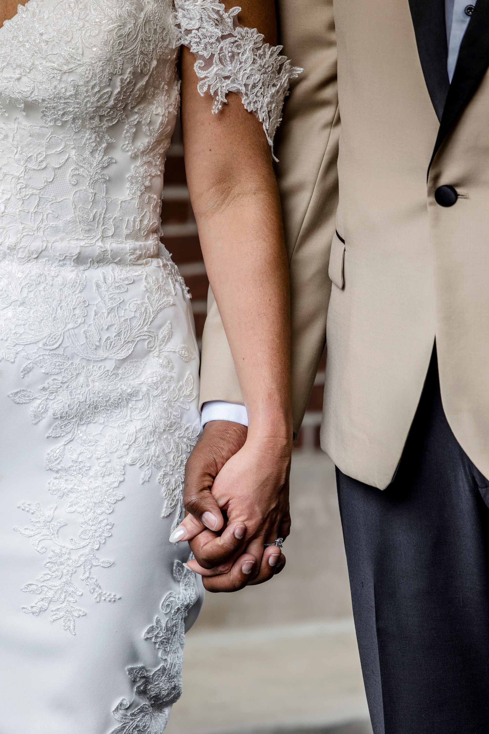 Bride and groom holding hands, with the bride in a white lace dress and groom in a tan jacket.