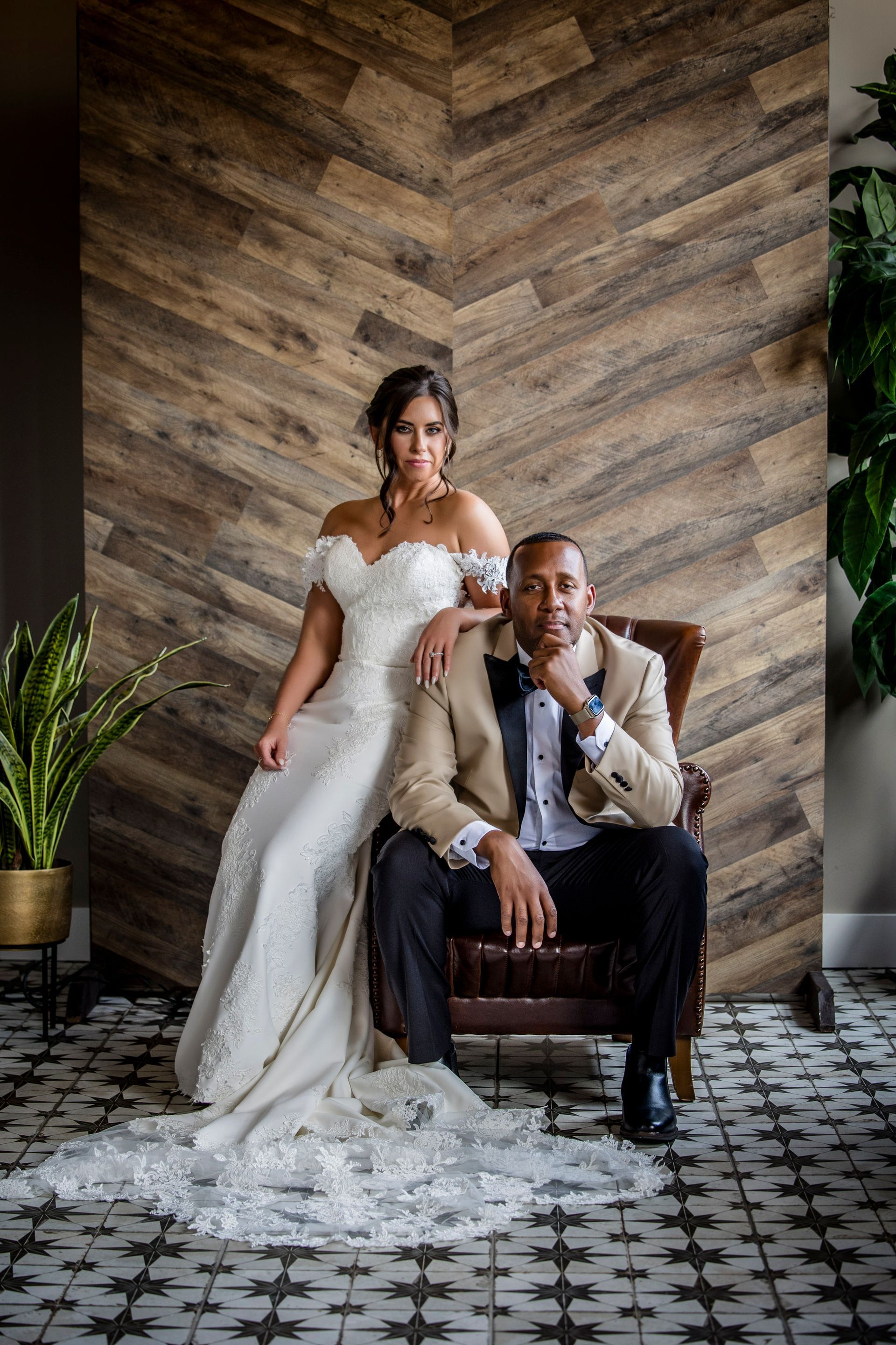 Two people in formal wear pose indoors against a herringbone wood wall, one standing and one seated.
