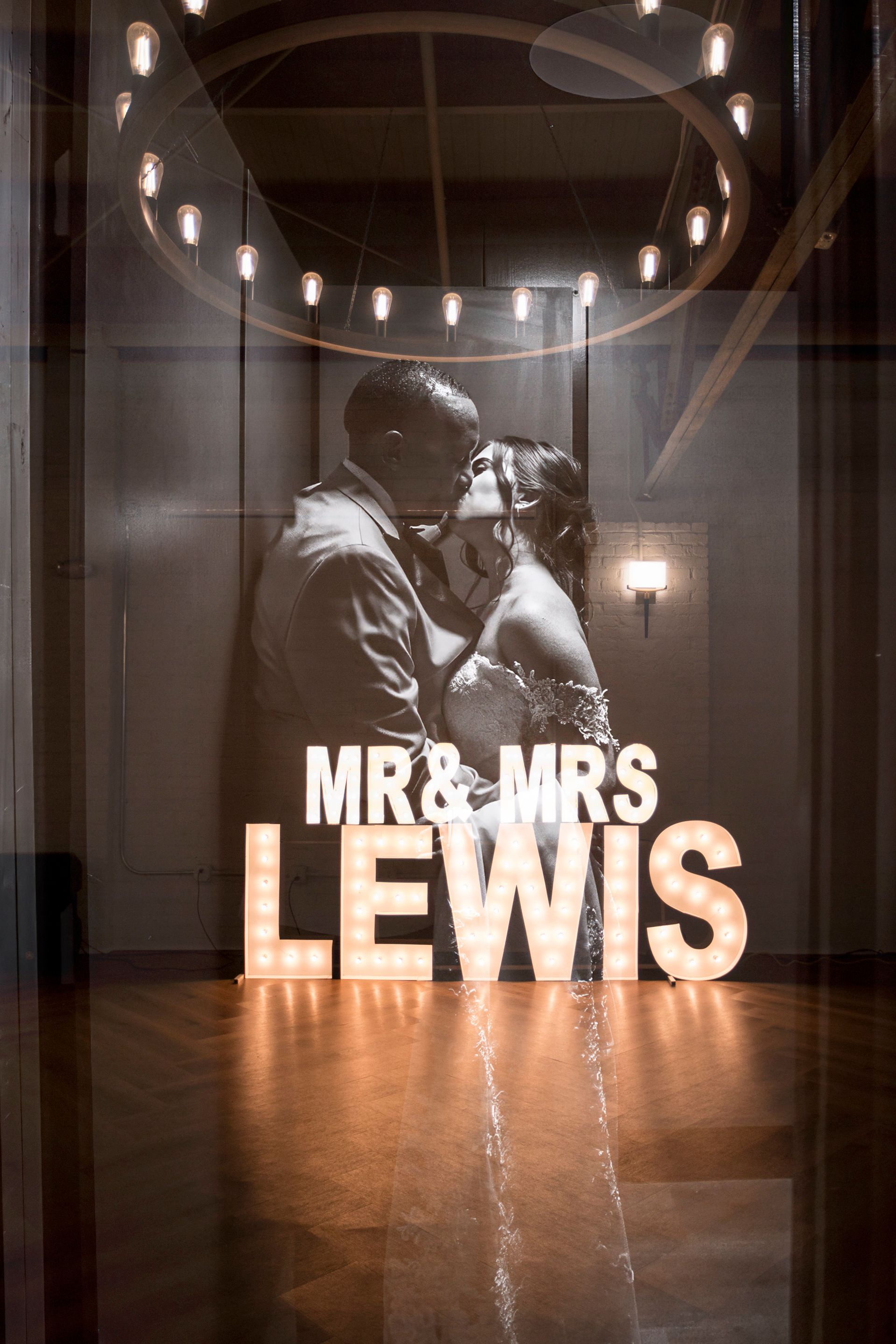 Couple embracing under “MR & MRS LEWIS” wedding sign in a warmly lit hallway
