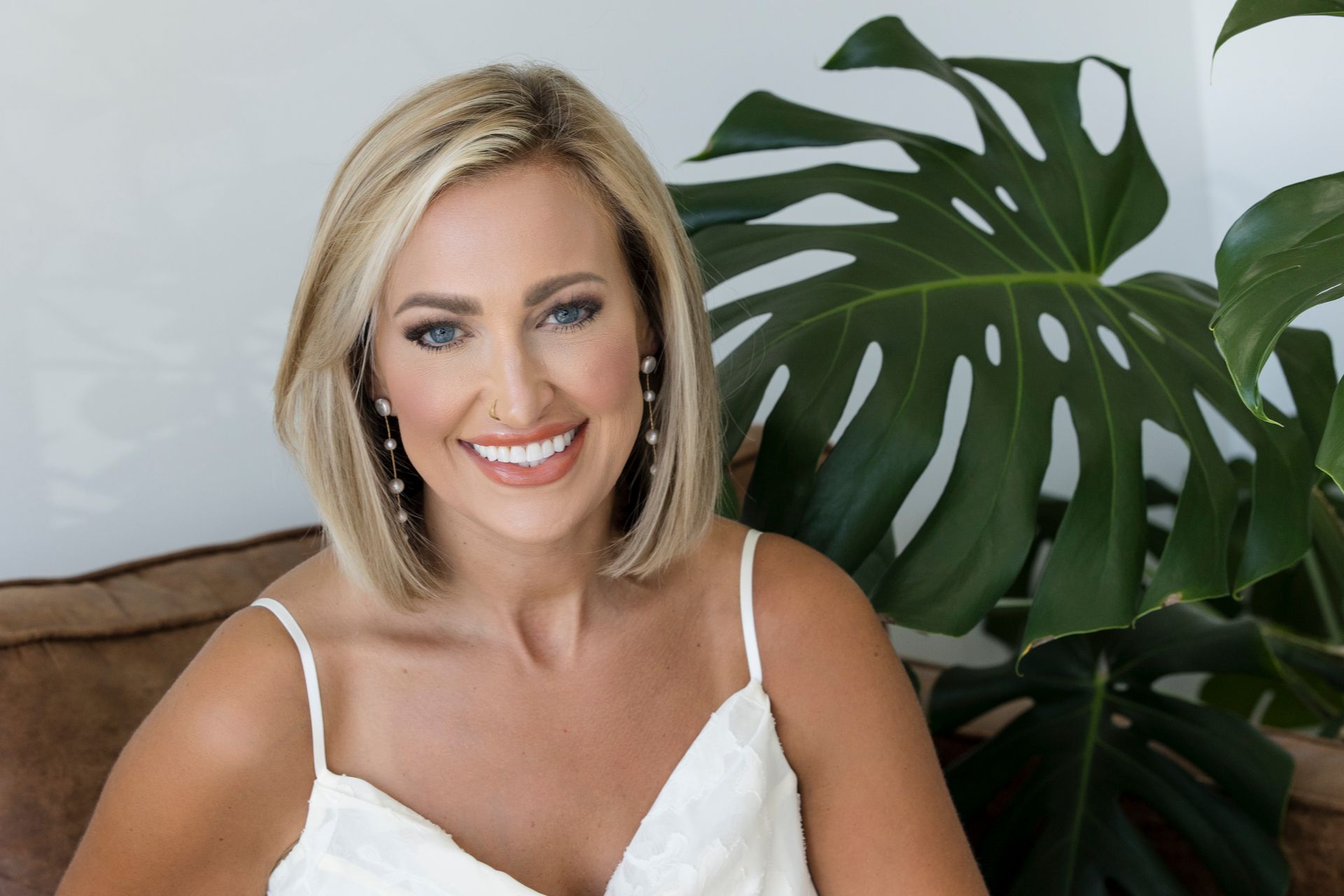 Smiling woman in a white top seated indoors beside a large green leaf plant