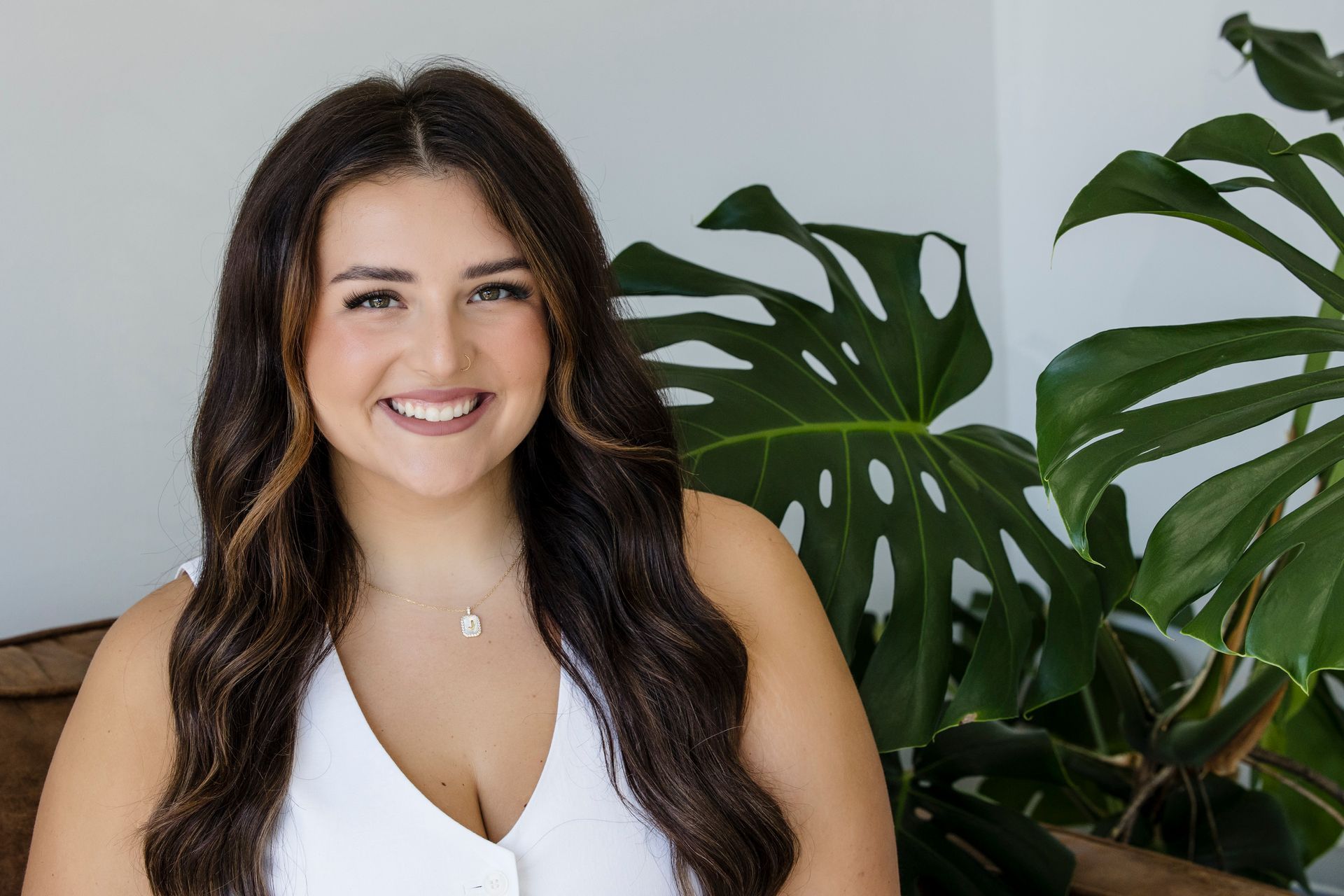 Smiling woman in a white top standing beside large green houseplants against a light wall
