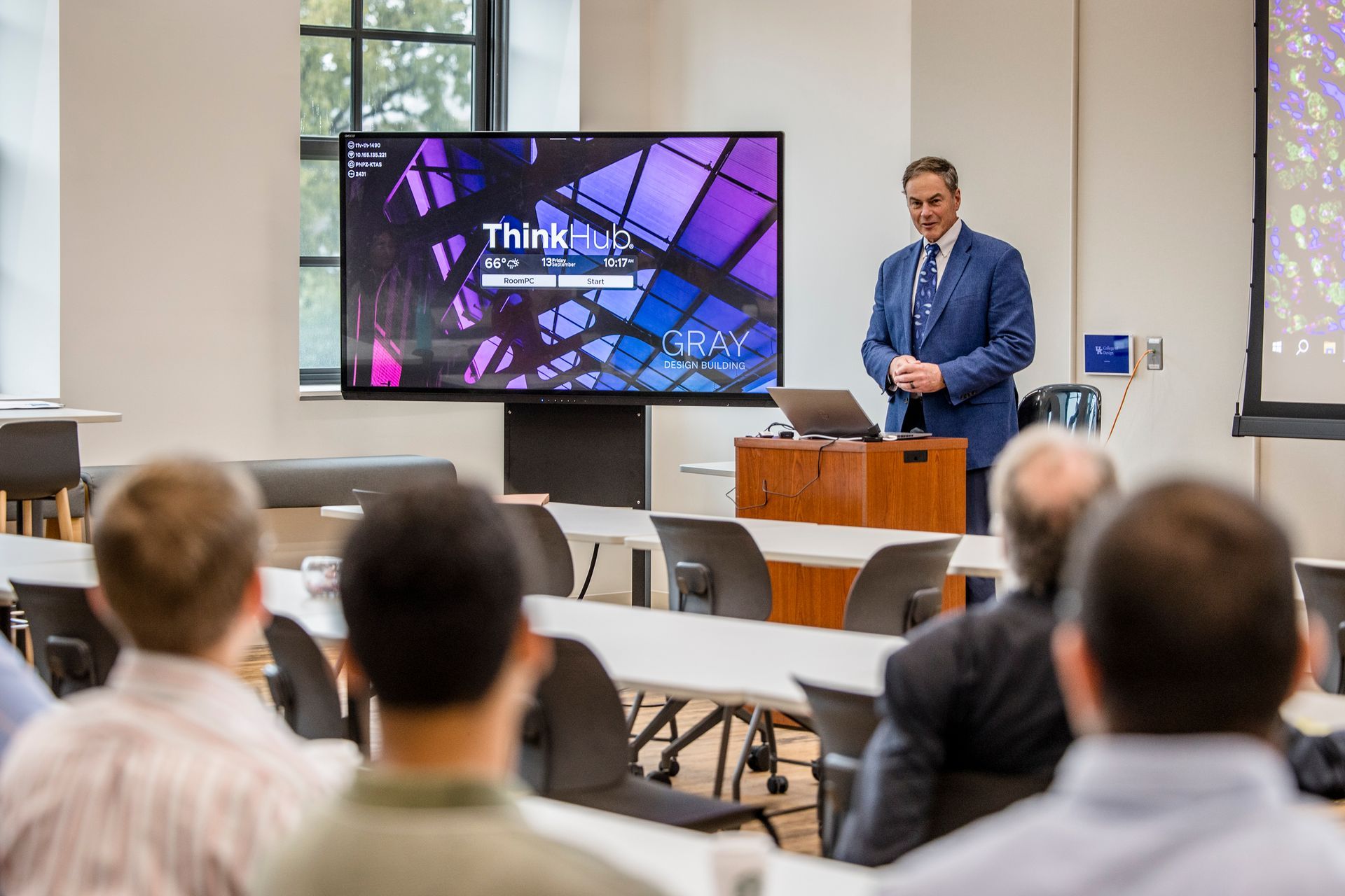 Speaker presenting in a conference room, with a slide reading “Think…” on a large screen.