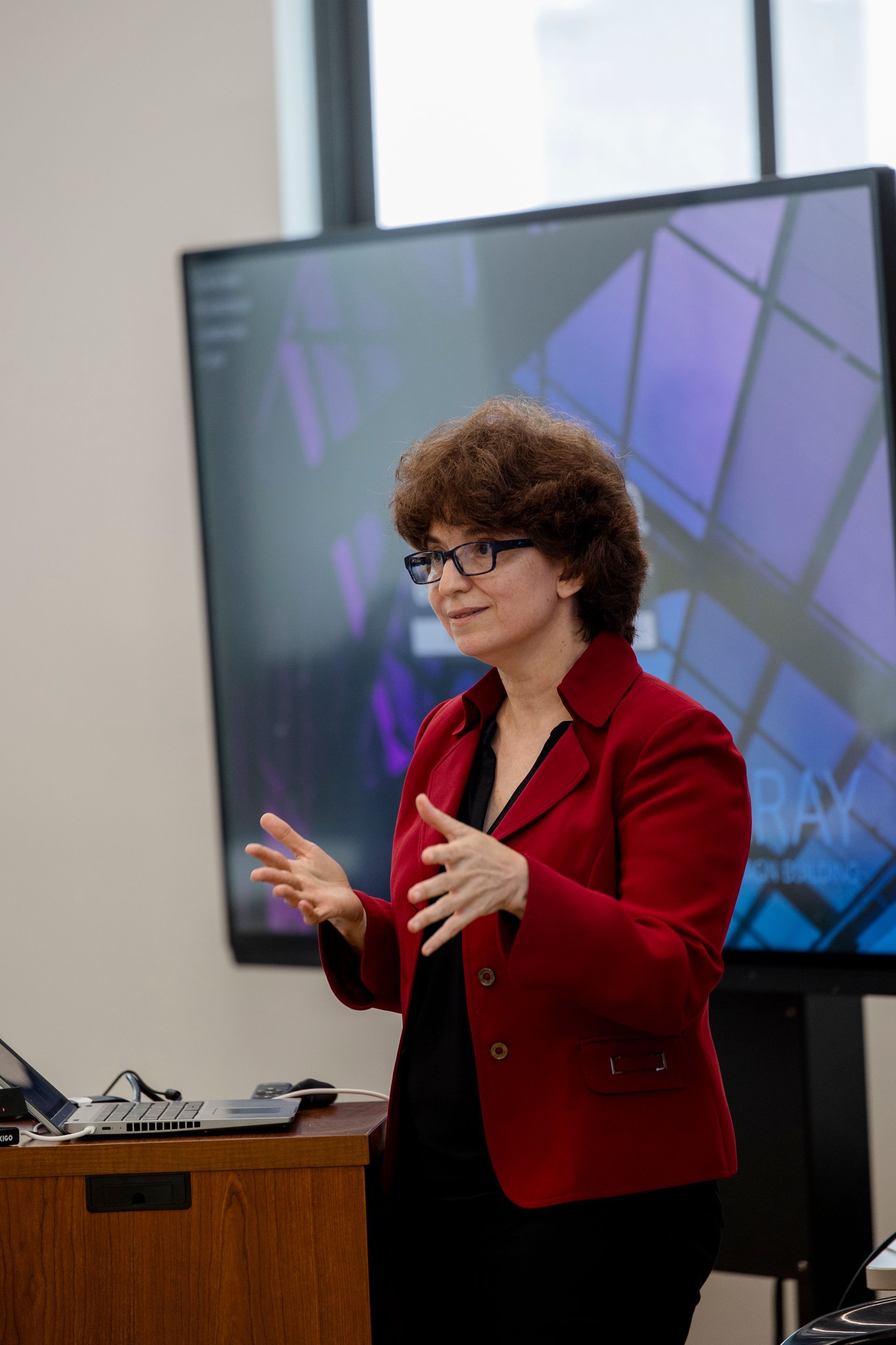 Woman in a red blazer speaking beside a large display, gesturing during a presentation.