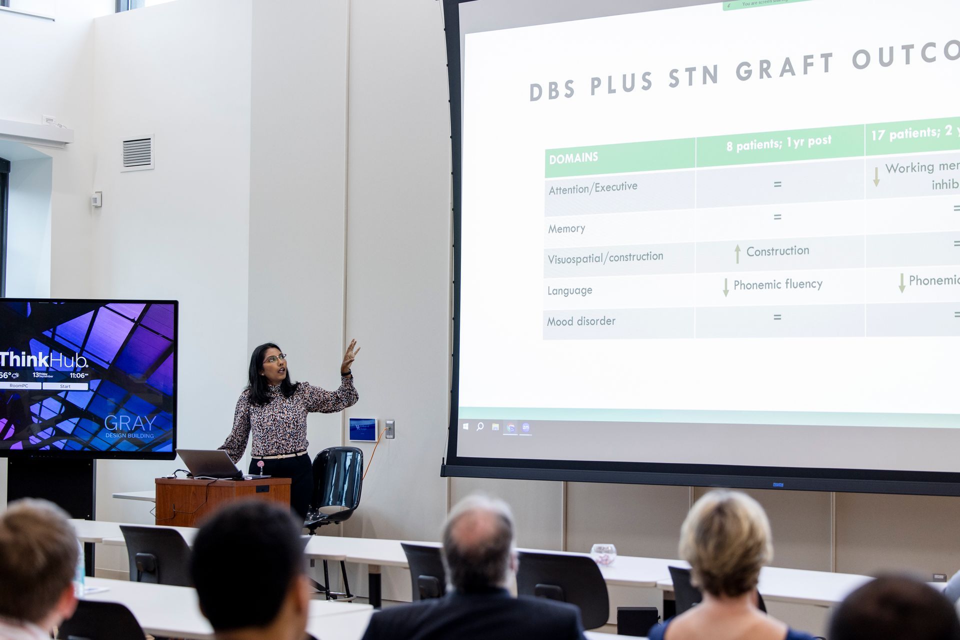 Presenter gestures beside projected slide in a conference room with seated audience.