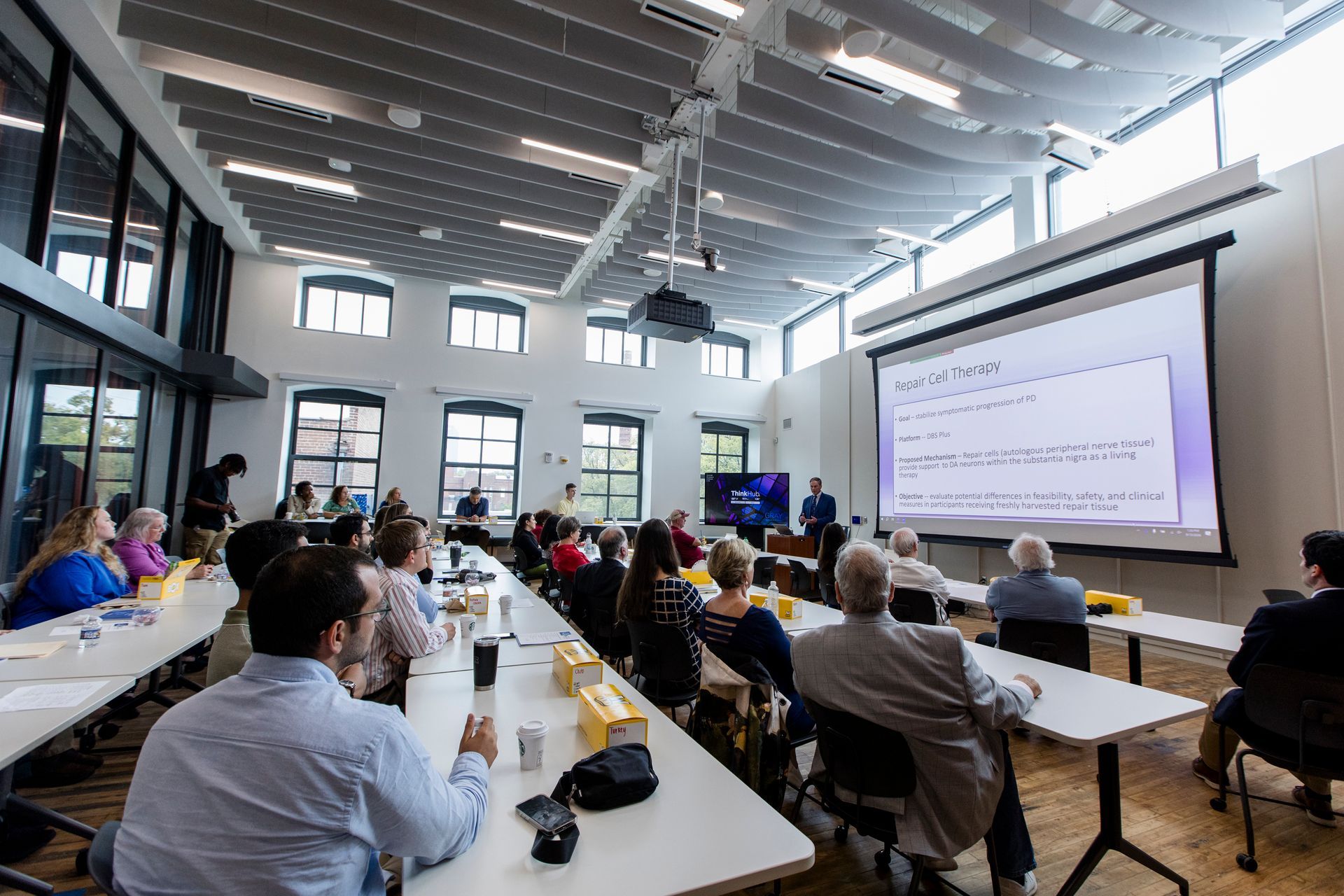 Audience in a bright conference hall watches a speaker present slides on a large screen.