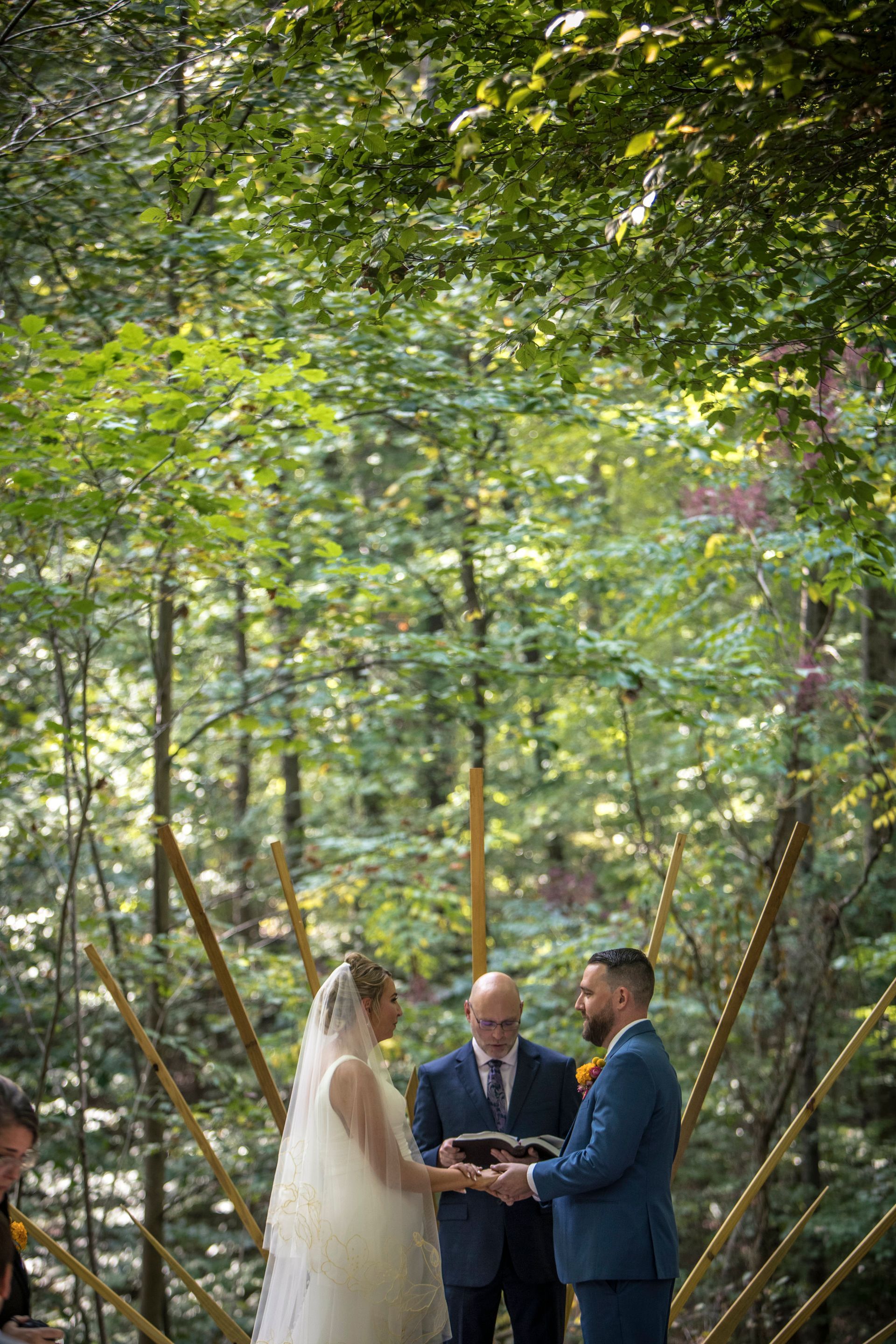 Outdoor wedding ceremony in a forest, couple exchanging vows under green trees with an officiant.