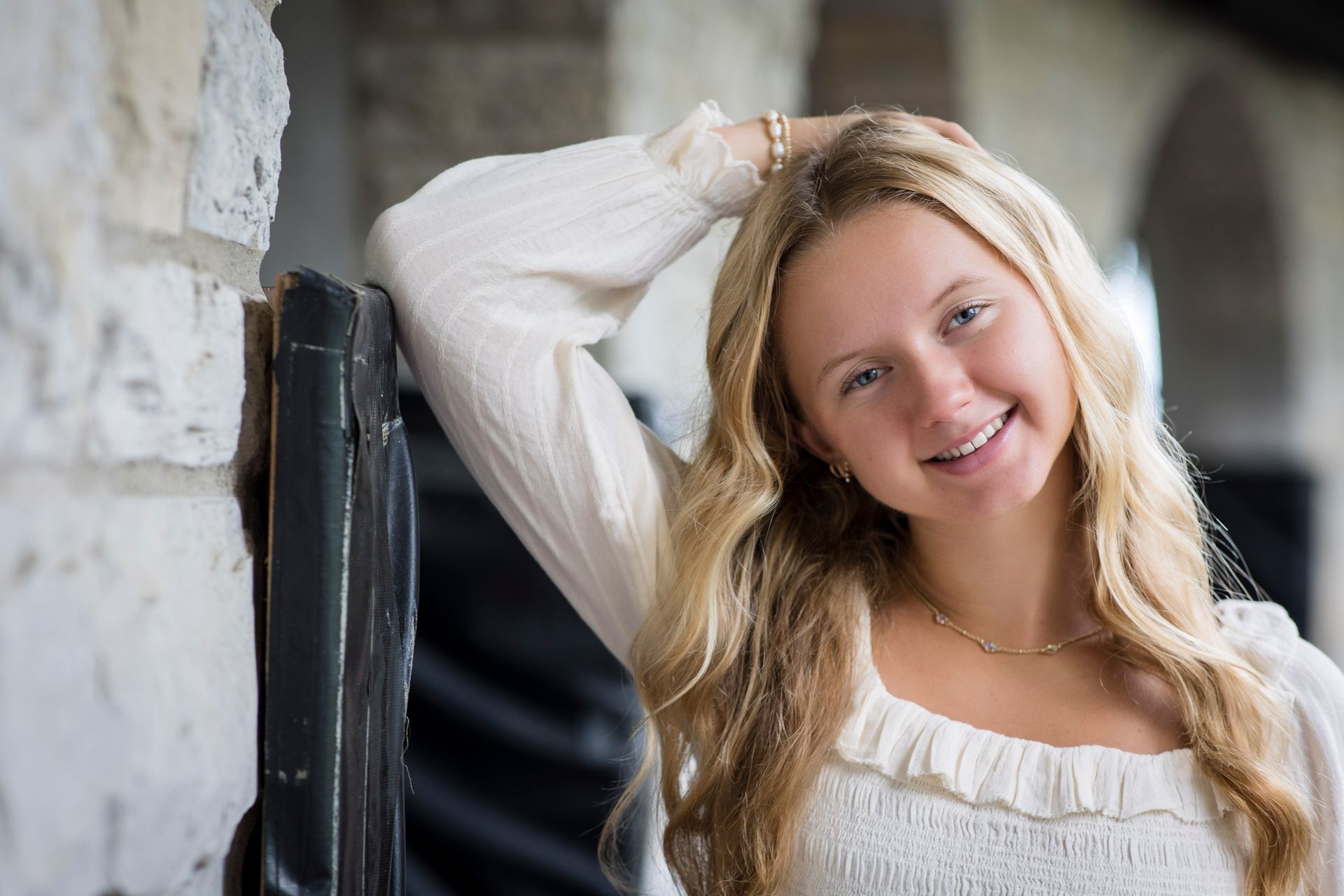 Smiling blonde woman in a white blouse leaning against a stone wall indoors