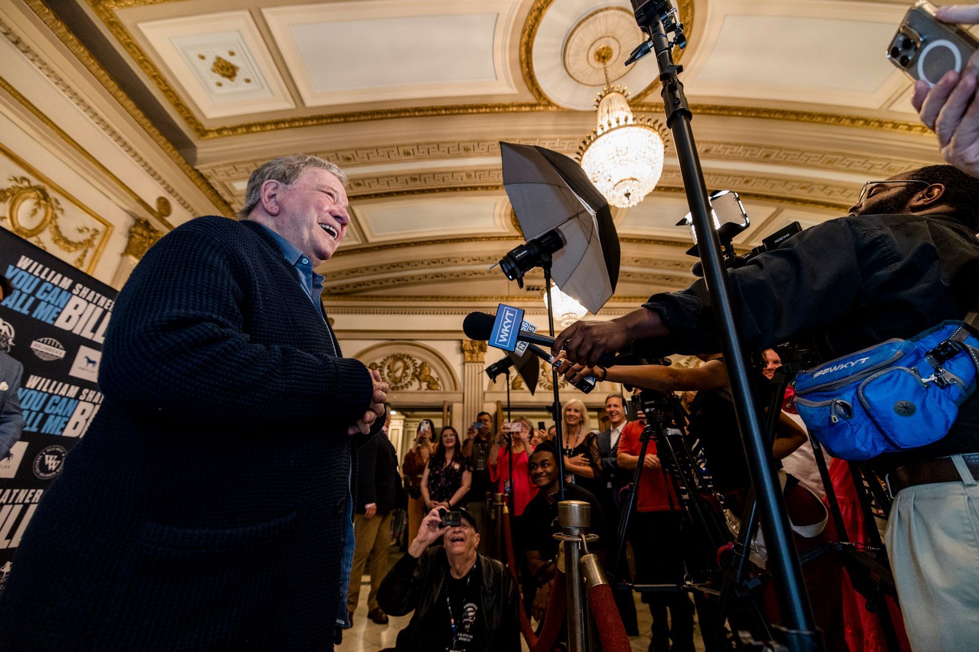 Man speaking to reporters in an ornate room, with cameras, microphones, and flashlights aimed at him.