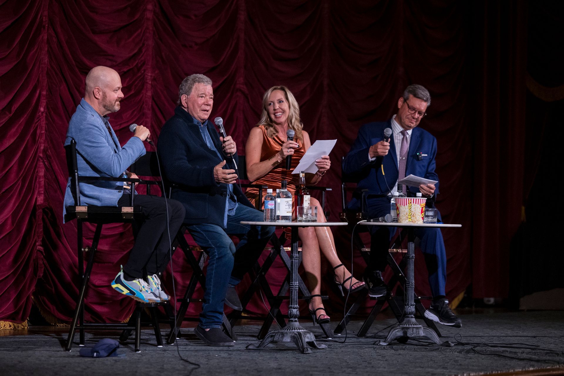 Four people on stools in a stage panel discussion, with microphones and papers before a red curtain.