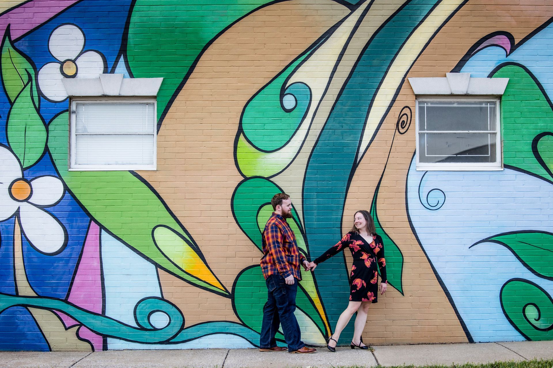 Two people stand in front of a colorful mural with flowing shapes and two windows.