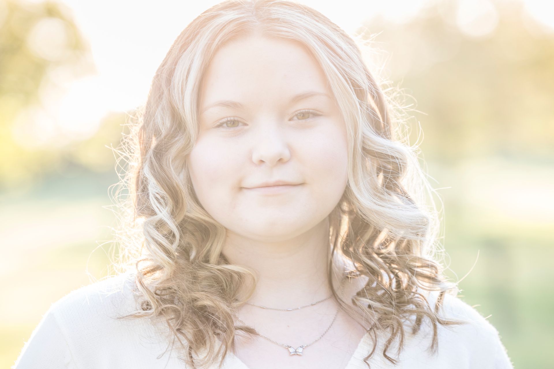 Young person with wavy blonde hair, softly lit outdoors, looking at the camera with a slight smile.