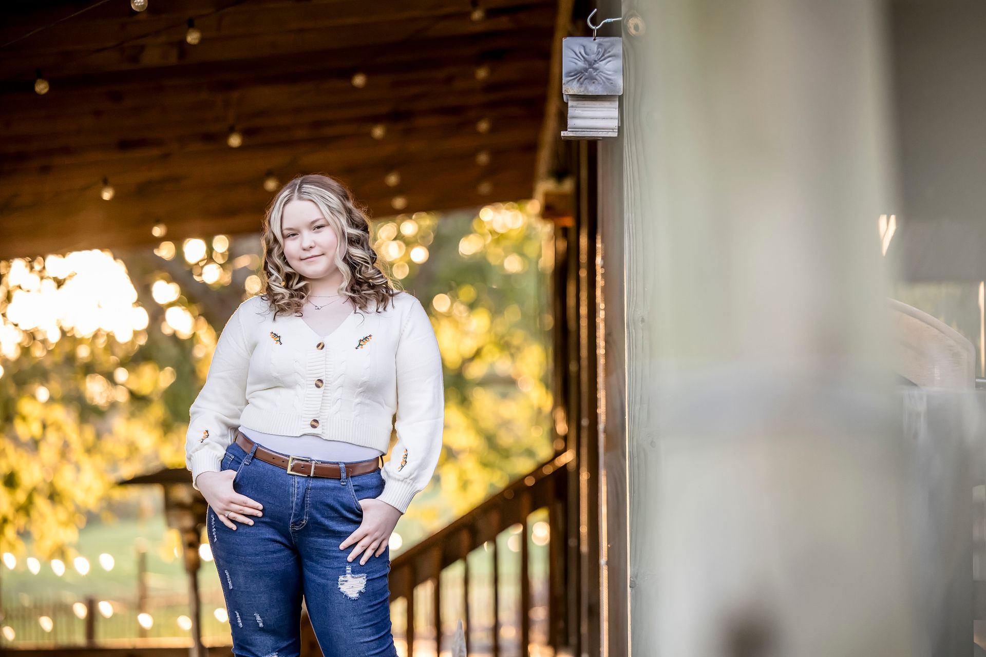 Woman in a white blouse and jeans standing on a porch with warm sunset lights in the background